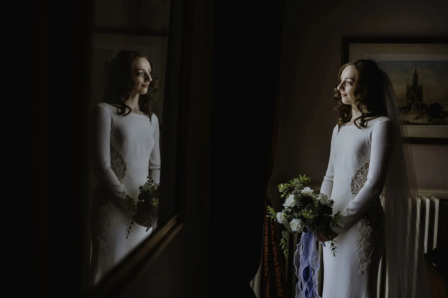 A bride in a white wedding gown with lace details, holding a bouquet, standing in front of a mirror, smiling gently, with her reflection visible.