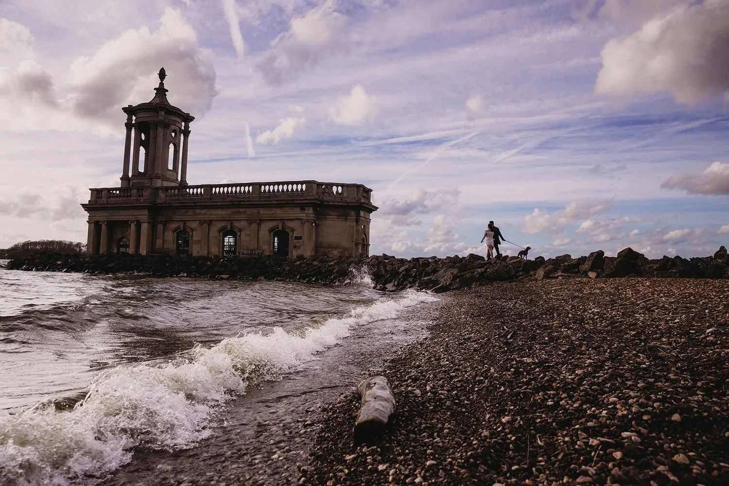 A person walking a dog along a rocky shoreline with an ornate stone building featuring a tower and balustrade in the background, under a partly cloudy sky.