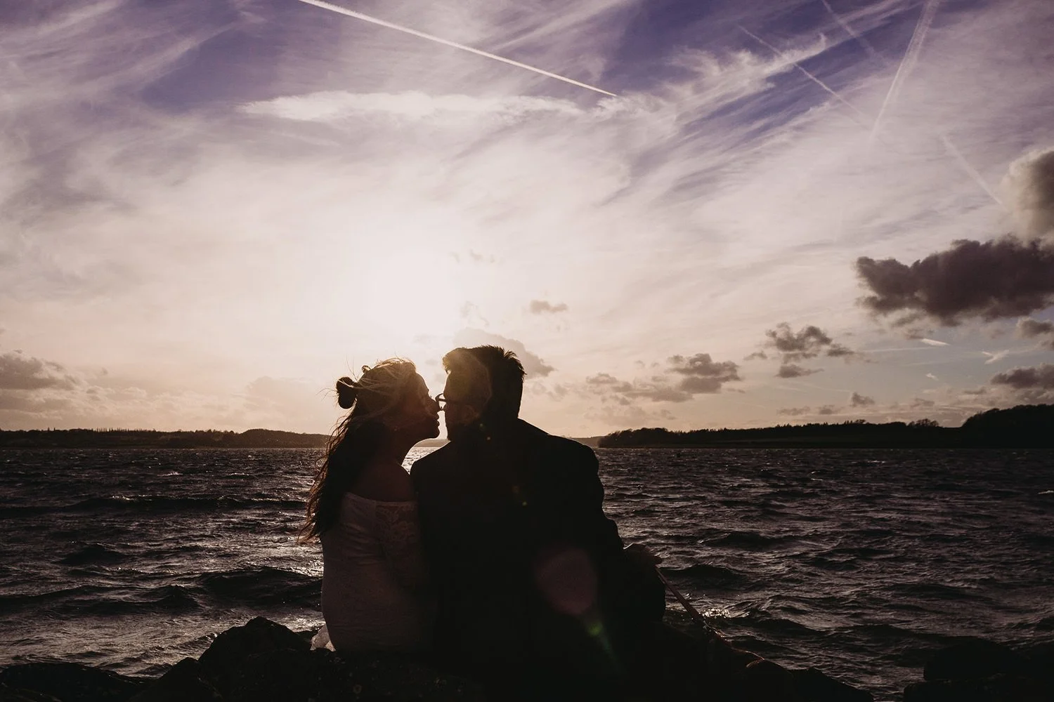 Silhouetted couple sitting on rocks by a body of water during sunset, with clouds and contrails in the sky.
