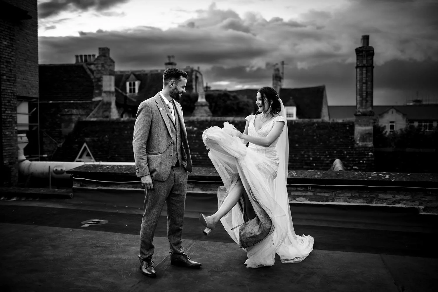 Black and white photo of a bride and groom on a rooftop with a cityscape background. The bride is playfully lifting her wedding dress while the groom watches smilefully.