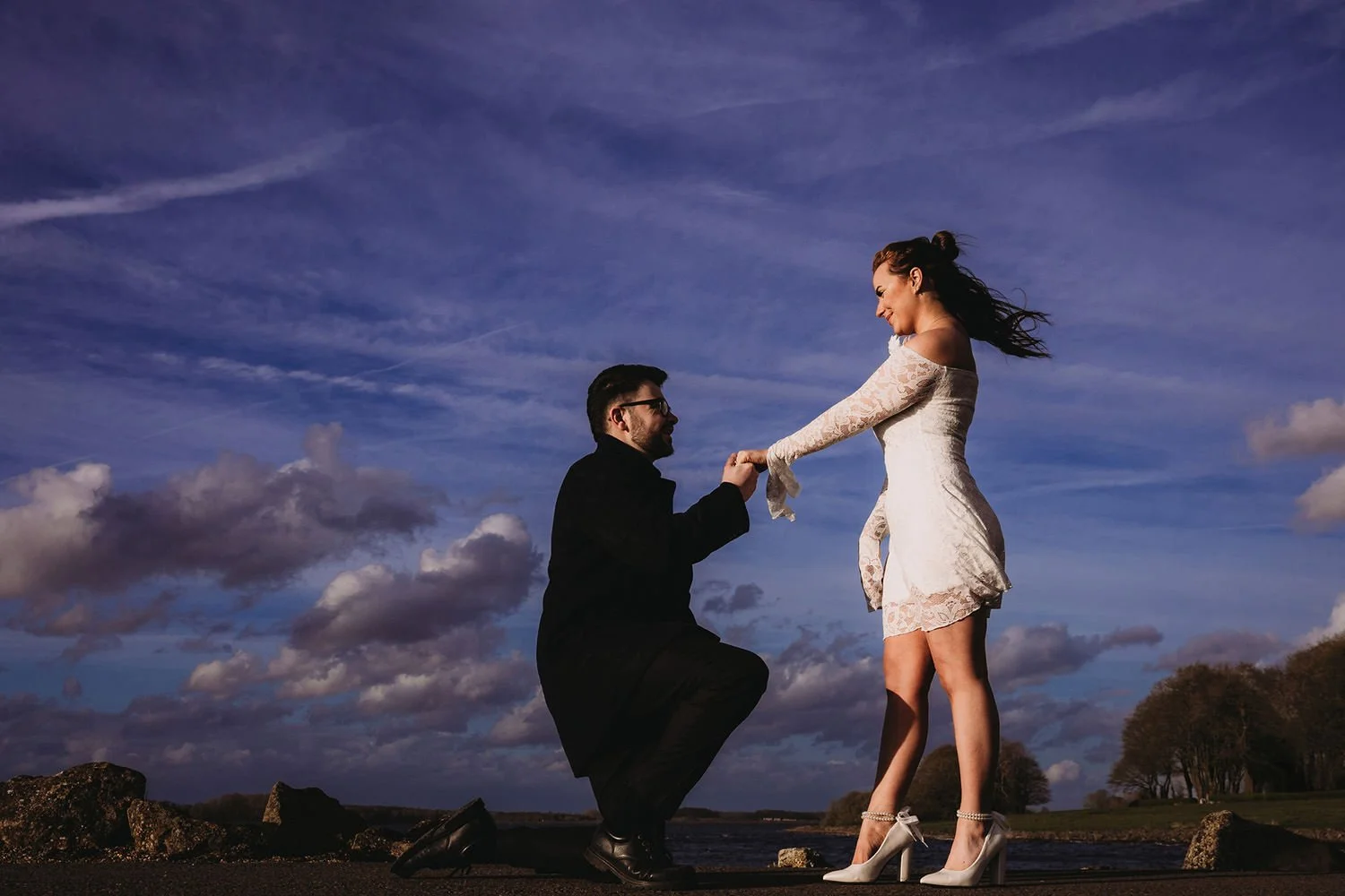 A man is proposing marriage to a woman outdoors, with a scenic sky and trees in the background. The woman is smiling and wearing a white lace dress and high heels, while the man is kneeling and holding her hand.