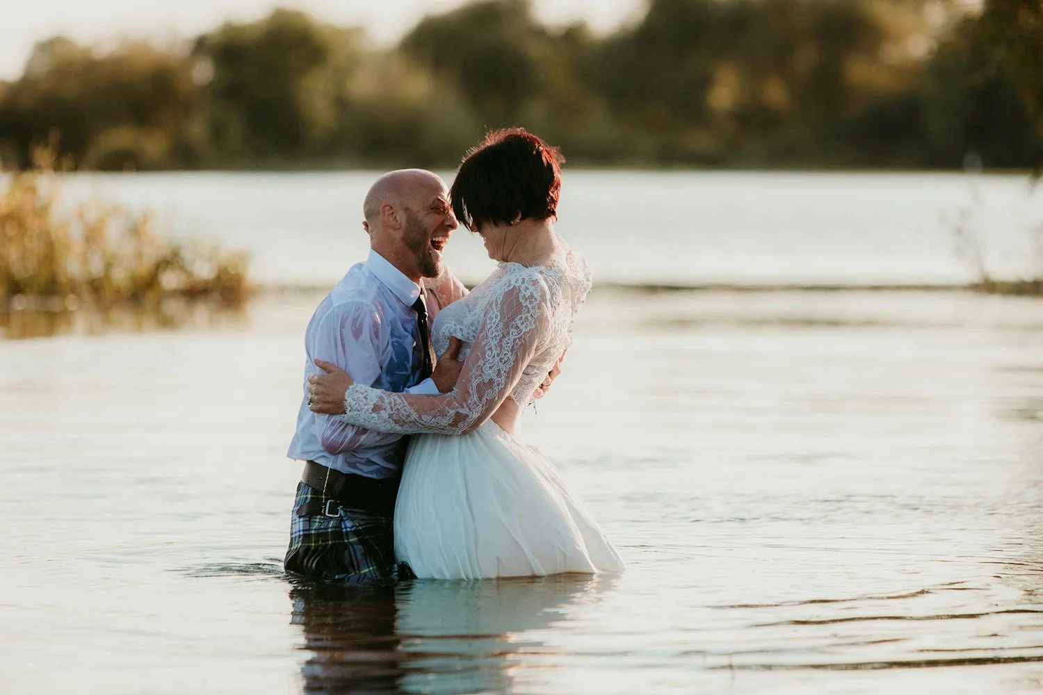 A couple in wedding attire, standing in a lake, happily laughing and embracing at sunset.