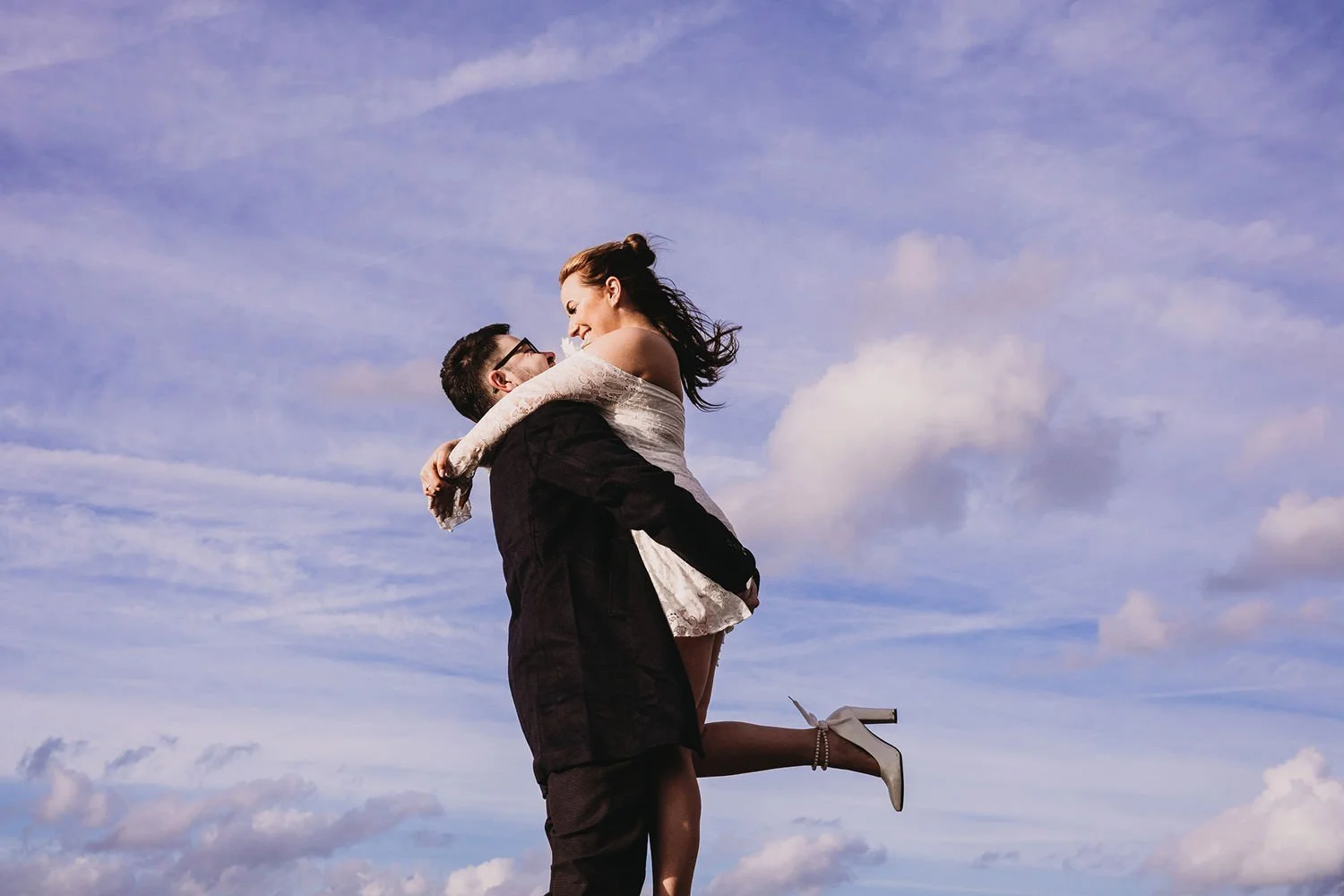 A man lifting a woman in a white dress and high heels into the air outdoors with a cloudy sky background.