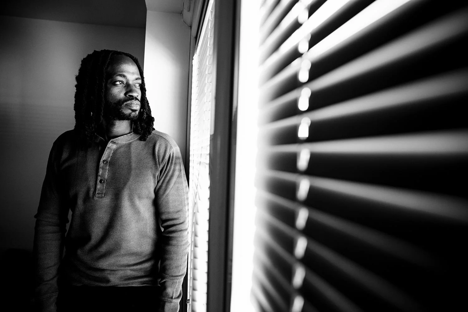 Black and white photo of a man with dreadlocks looking out a window with blinds.