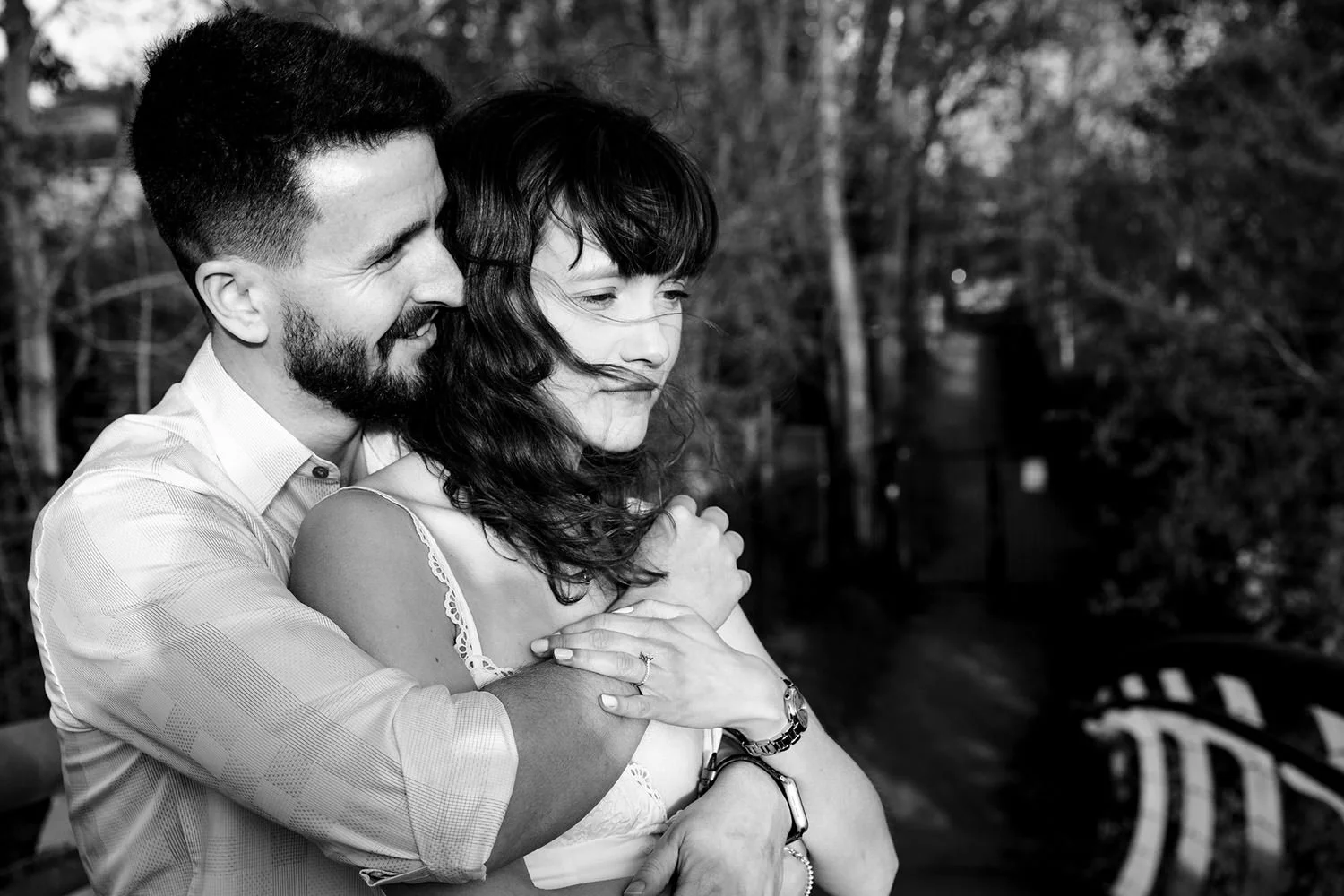 A black and white photo of a couple embracing outdoors. The man is smiling and has a beard, the woman has long hair and is looking down with a gentle expression. Trees and a pathway are in the background.