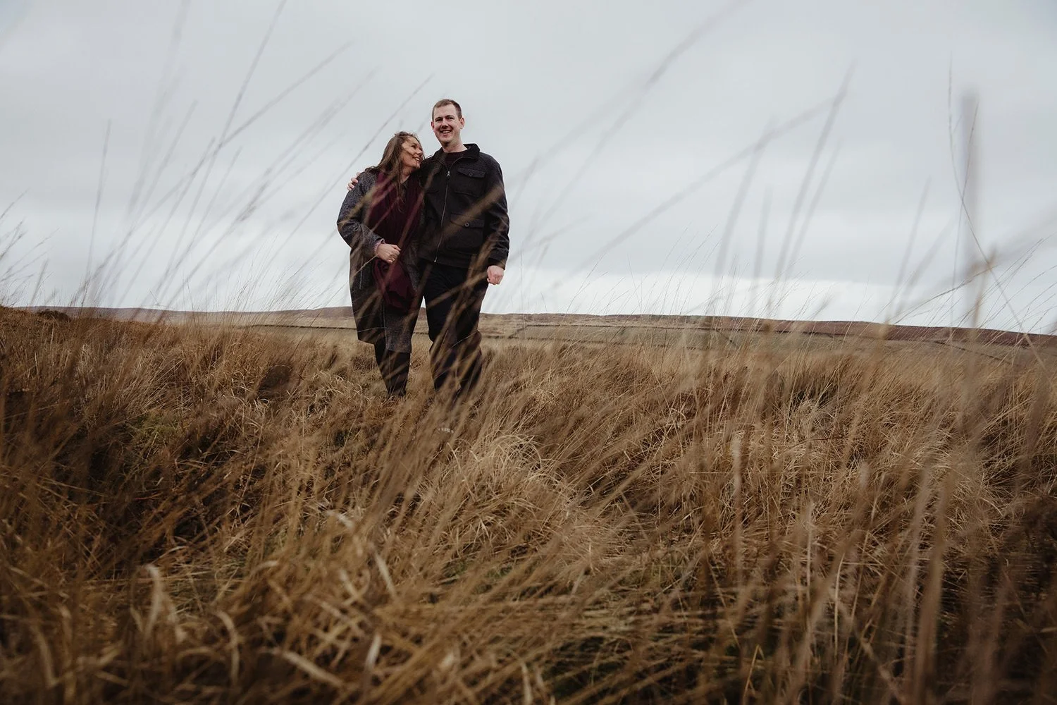 A couple walking and smiling in a field of tall, golden grass on an overcast day.