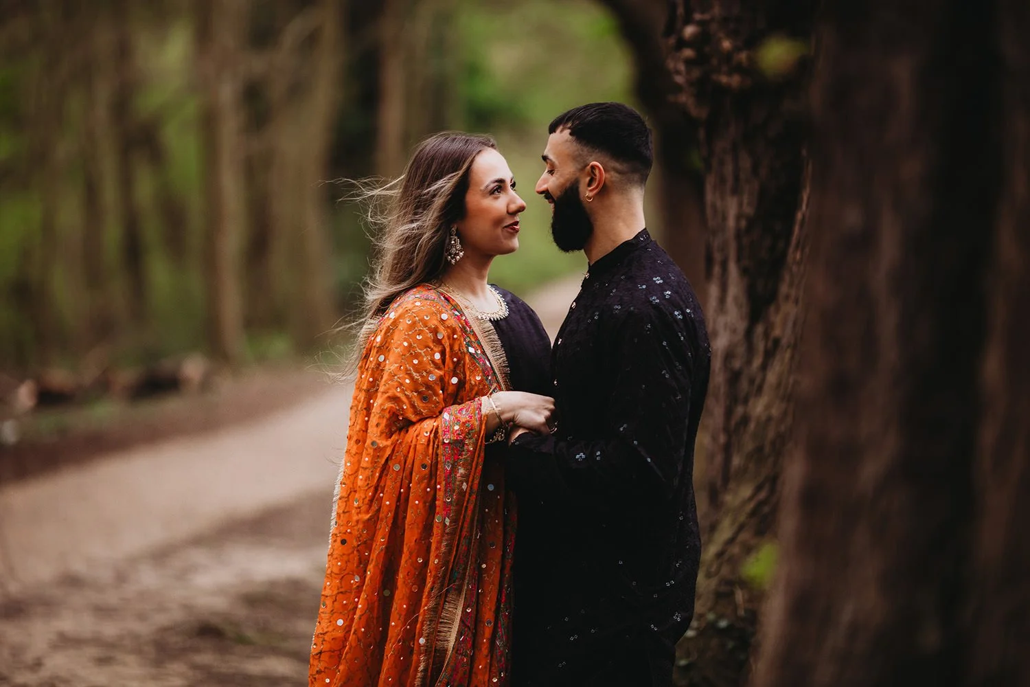 Engaged couple standing close together on a forest path, gazing into each other's eyes, with trees in the background. The woman wears an orange traditional garment with embellishments, and the man wears a black patterned outfit.