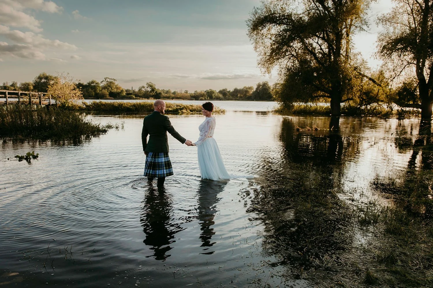 A bride and groom stand in a river holding hands at sunset, with trees and a bridge in the background.