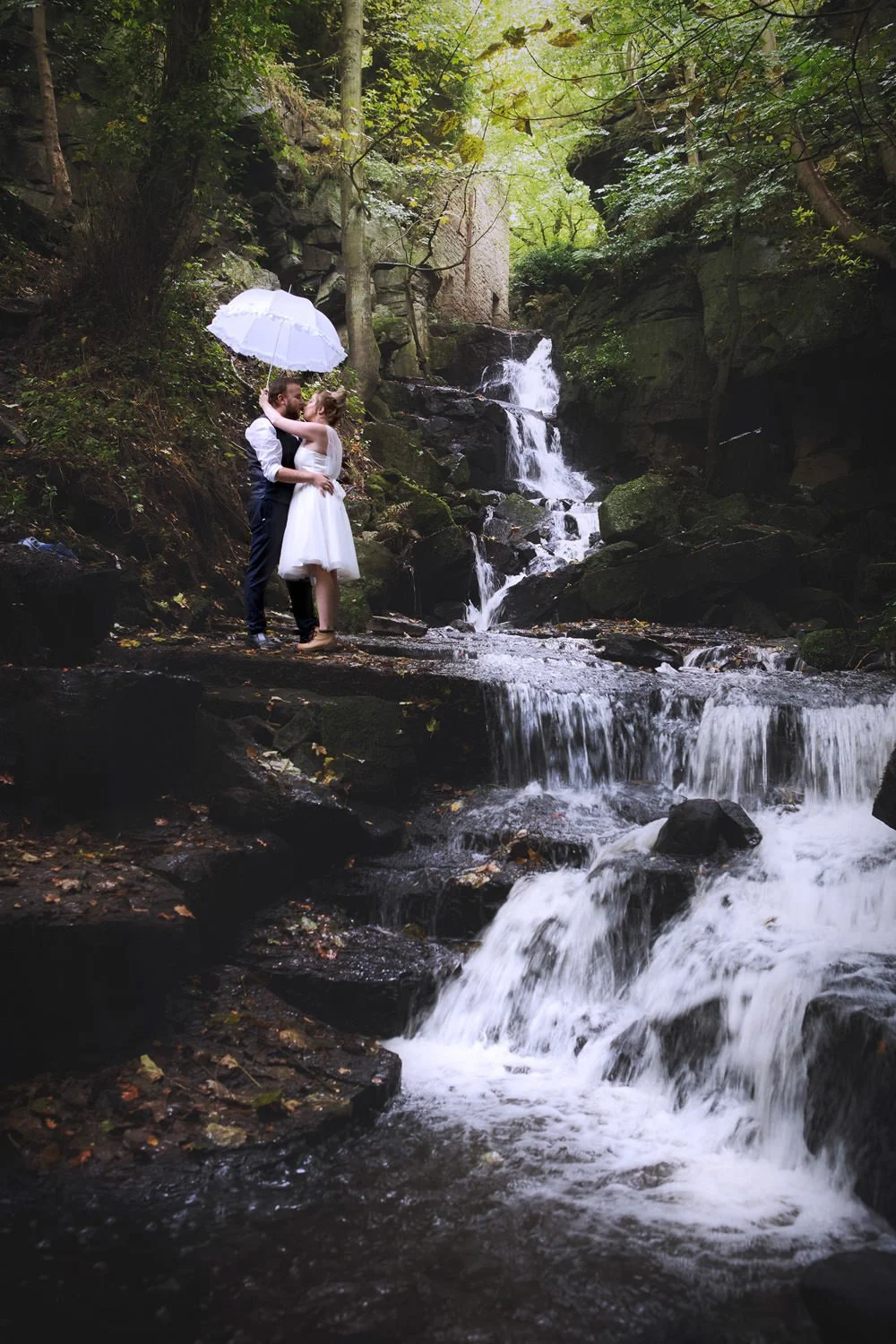 A couple in wedding attire stands embracing under a white umbrella by a waterfall in a forested area.