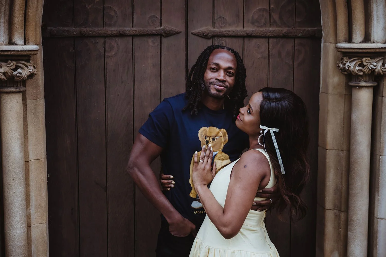 A man and woman standing close together in front of a wooden door. The man is wearing a navy blue T-shirt with a bear graphic and has shoulder-length dreadlocks. The woman is wearing a sleeveless yellow dress and has long dark hair styled in loose wa