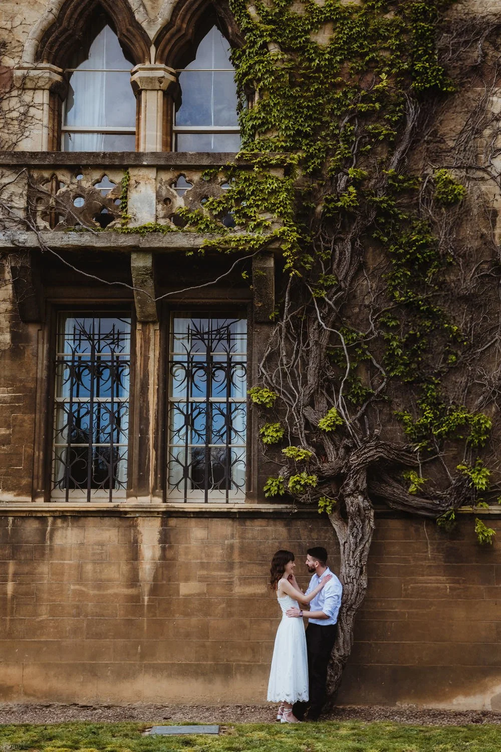 Engaged couple in Oxford standing close together under a large, old, gnarled tree in front of a historic stone building with stained-glass and barred windows.