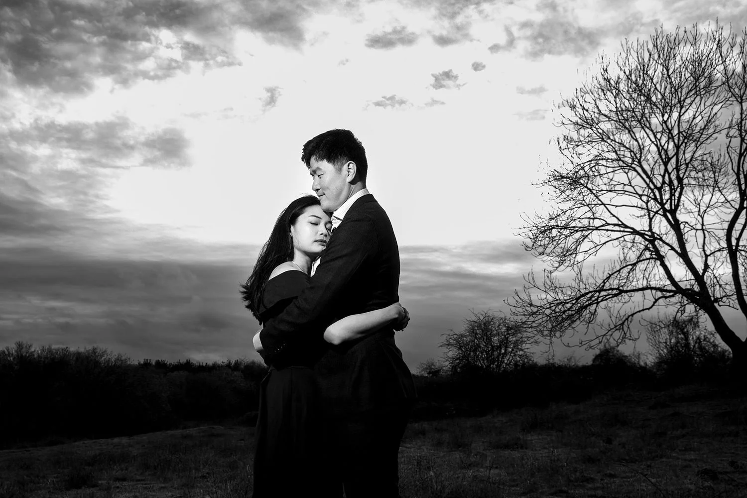 A black and white photo of a couple embracing outdoors at sunset. The woman has her eyes closed, and the man looks down at her, both with a peaceful expression. Bare trees are visible in the background.