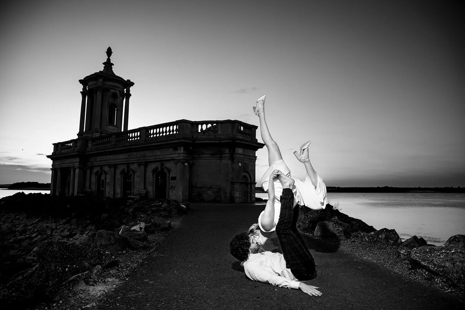 A couple performing acro yoga on a rocky path near water at sunset, with historic building in background.