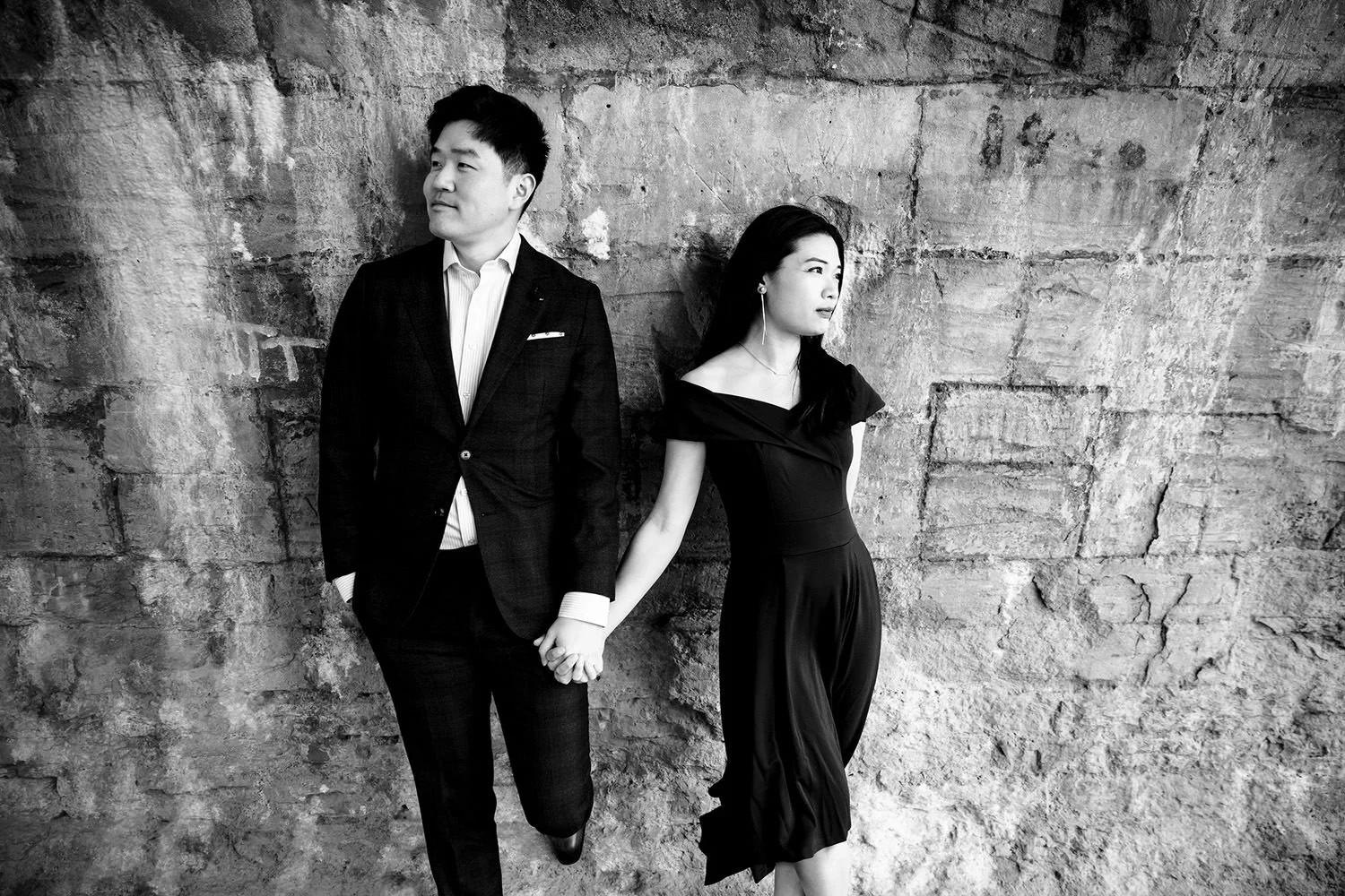 A black-and-white photo of a man and woman standing against a textured brick wall, holding hands, with the man looking to the side and the woman looking forward.