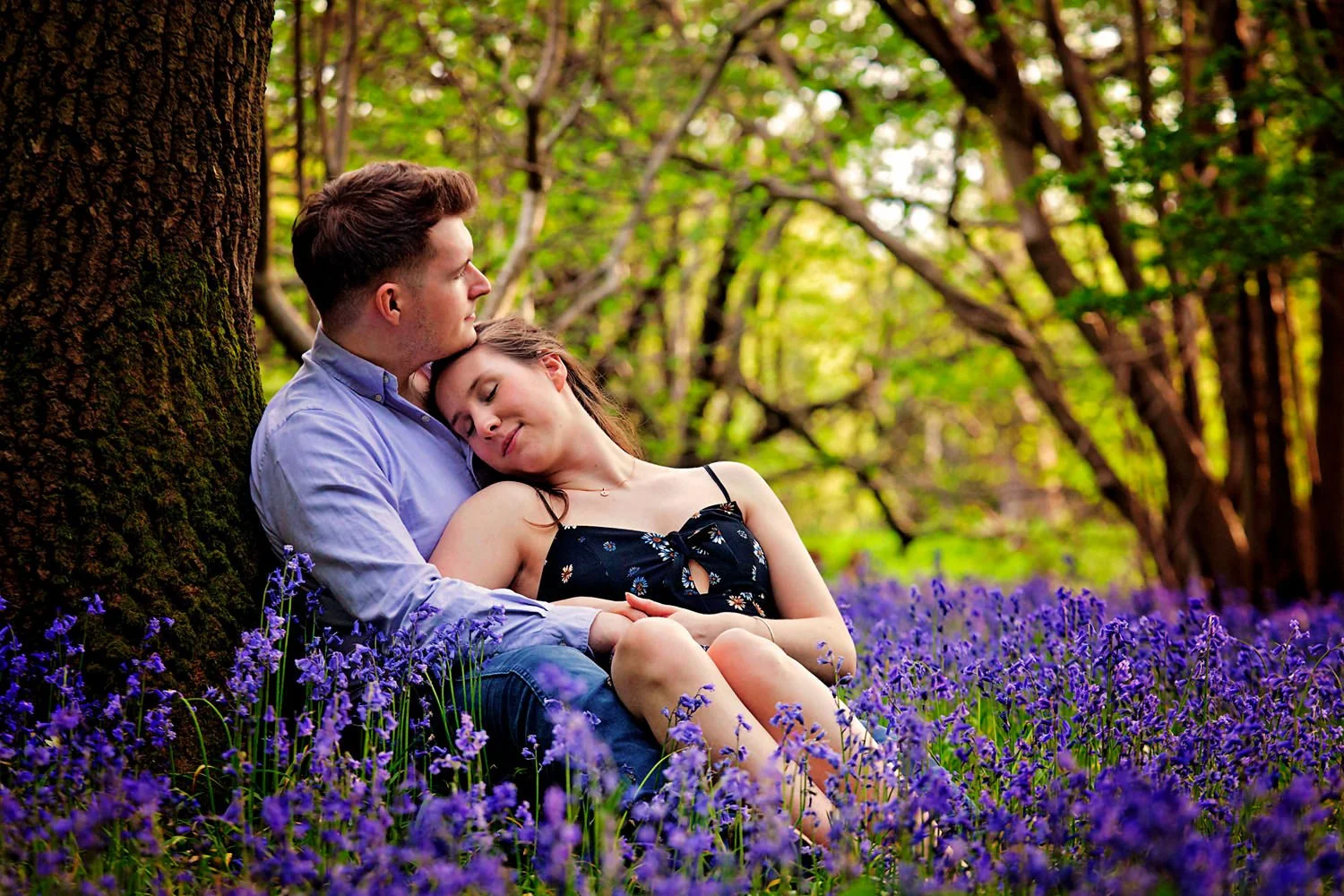 Engaged couple sitting under a tree in a forest, surrounded by bluebell flowers. The woman is resting her head on the man's chest, both appearing peaceful and affectionate.