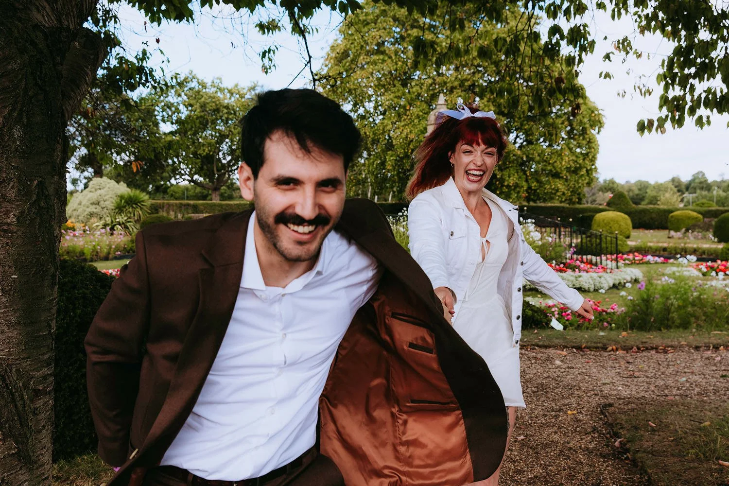 A man and woman smiling and holding hands while walking through a park with colorful flowers and trees in the background.