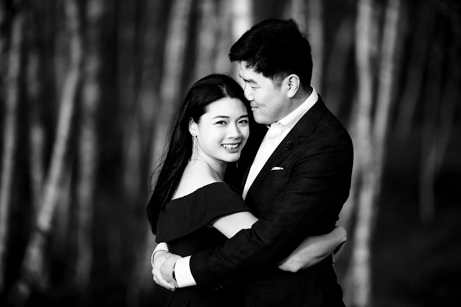 A black and white photo of a smiling woman and a man embracing, with a blurred background of trees.