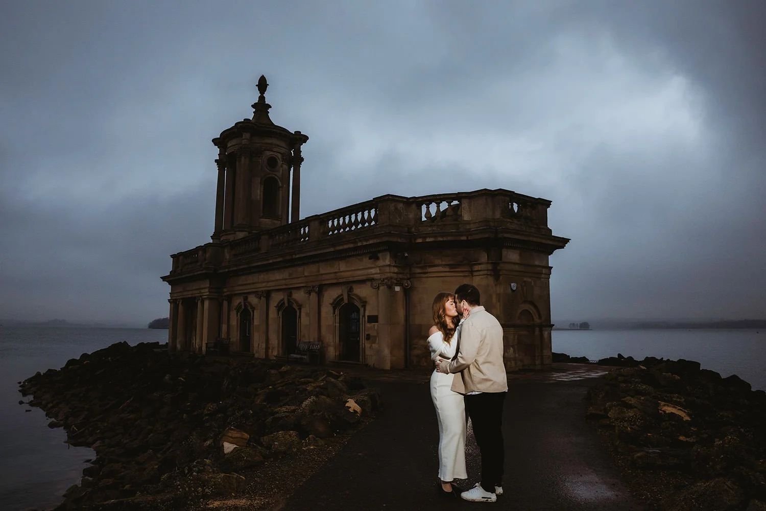 A couple in formal attire stands close and kisses on a path near a historic building by the water on a cloudy day.
