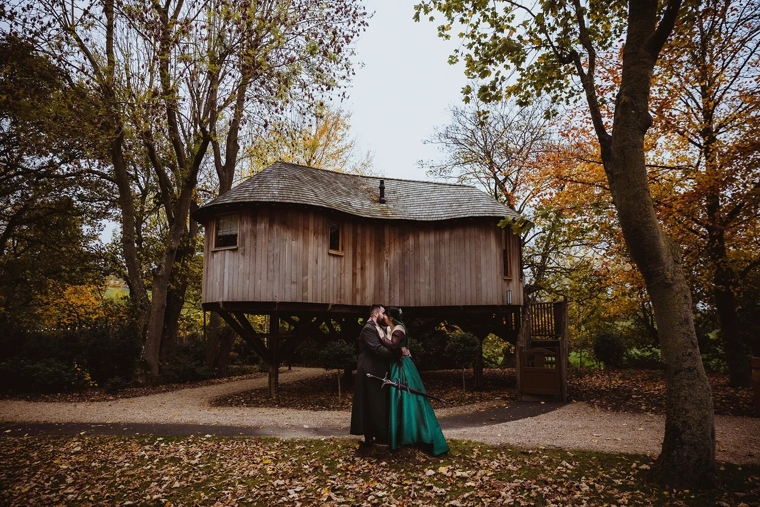 A couple dressed as a knight and a princess sharing a kiss in front of a treehouse in a park during autumn, with fallen leaves on the ground and colorful fall foliage on the trees.