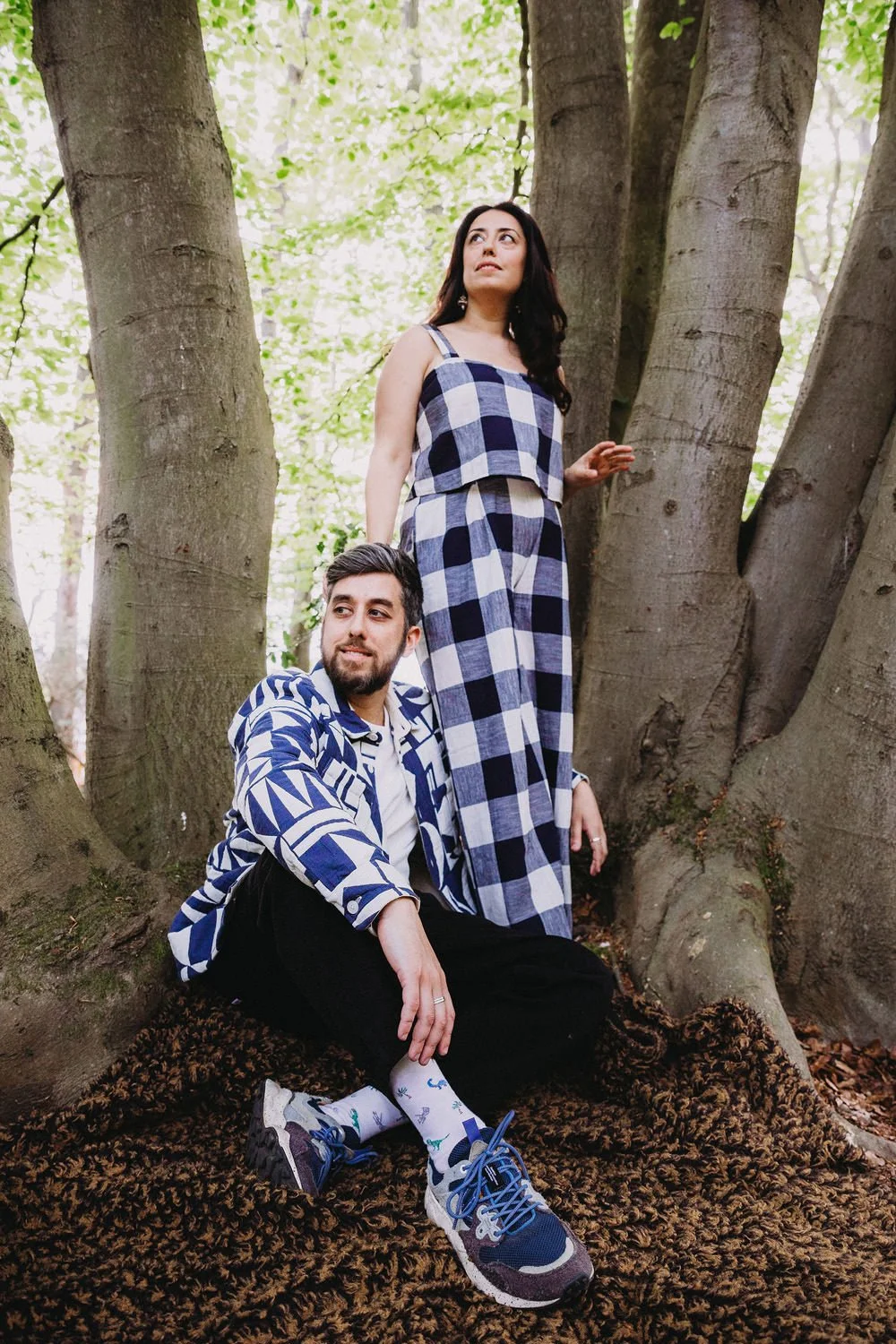 A woman standing on a large tree root and a man sitting on the ground in front of her, both wearing matching blue and white checkered outfits, in a lush green forest.