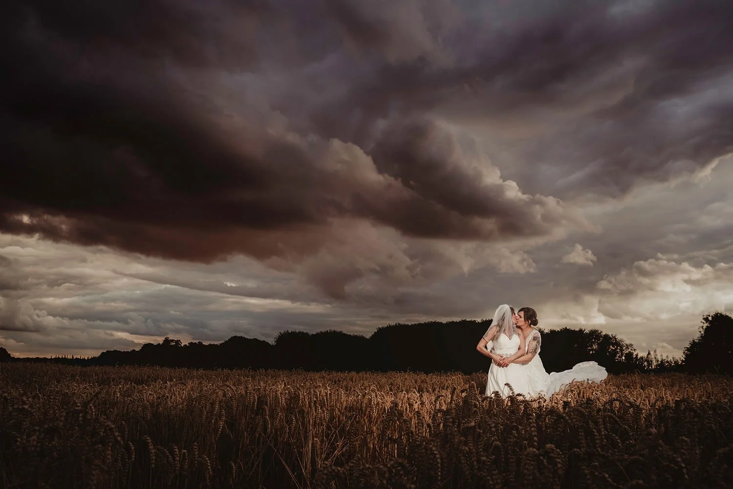 Two women dressed in wedding gowns sharing a kiss in a wheat field under a dark, cloudy sky.