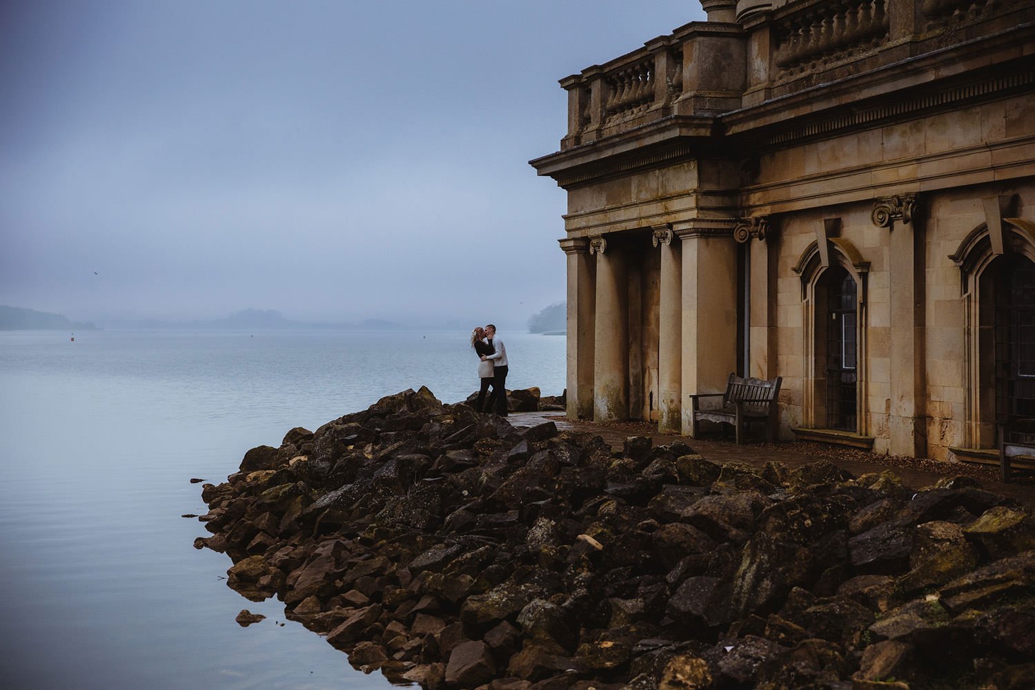A couple embracing on a rocky shoreline in front of an old building by the water on a foggy day.