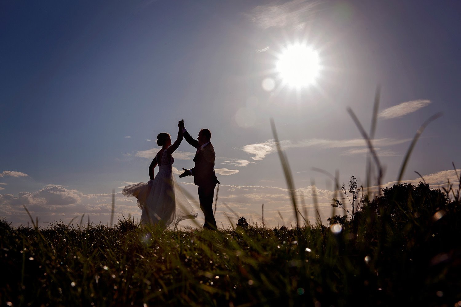 Silhouettes of a bride and groom dancing outdoors at sunset, with grass in the foreground and the sun shining brightly in the sky.