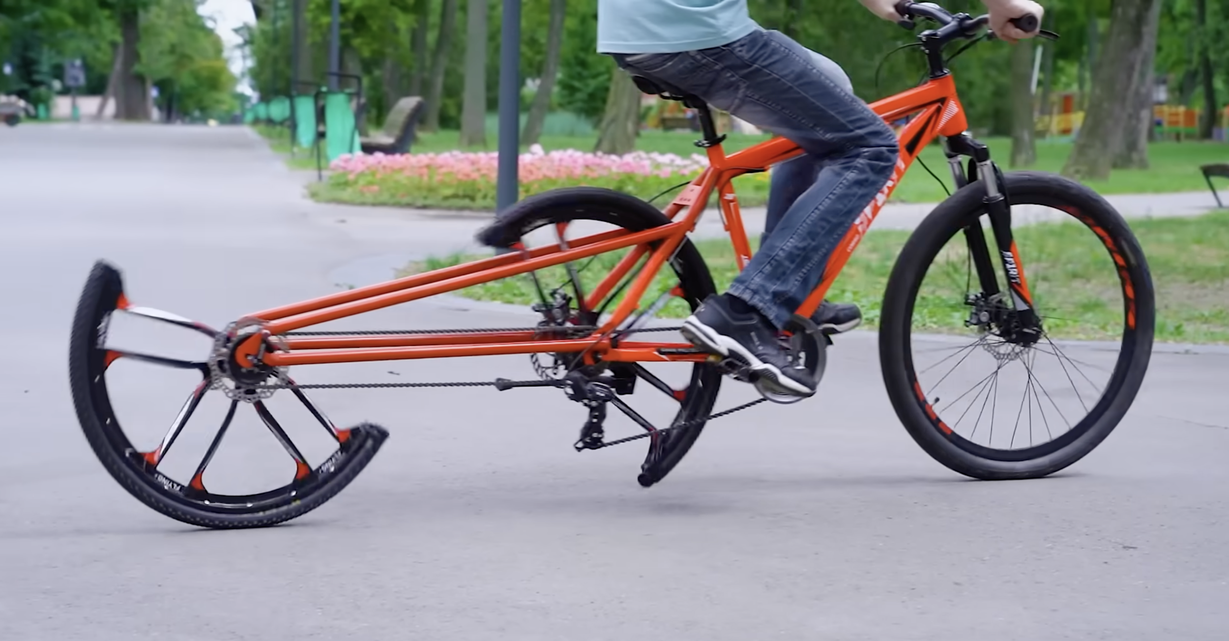Man rides red bicycle with split rear wheel through park.