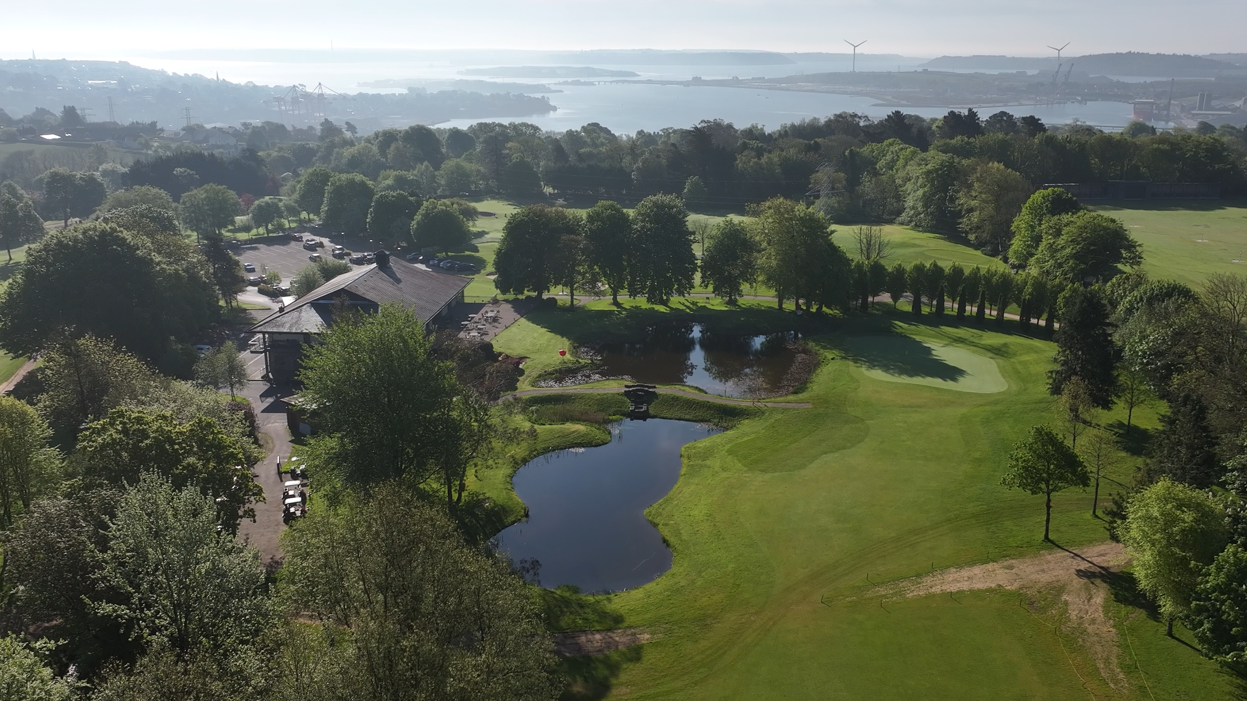 Aerial view of a golf course with green fairways, sand traps, small water hazards, surrounded by trees and a clubhouse, with a parking lot nearby, and a body of water and wind turbines in the distance.