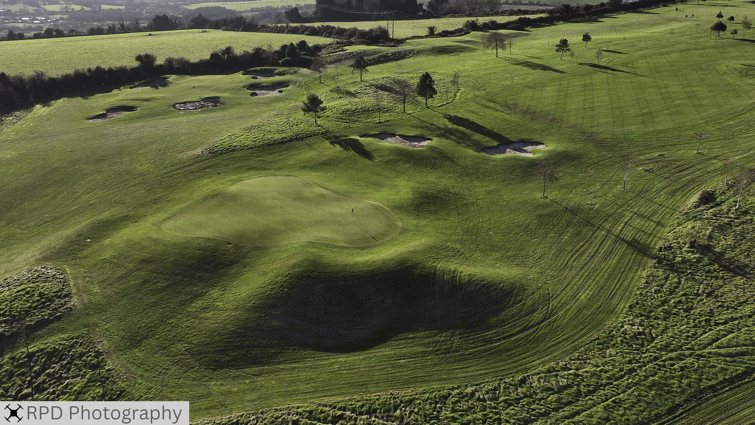 Aerial view of a golf course with greens, sand bunkers, and trees on rolling hills.