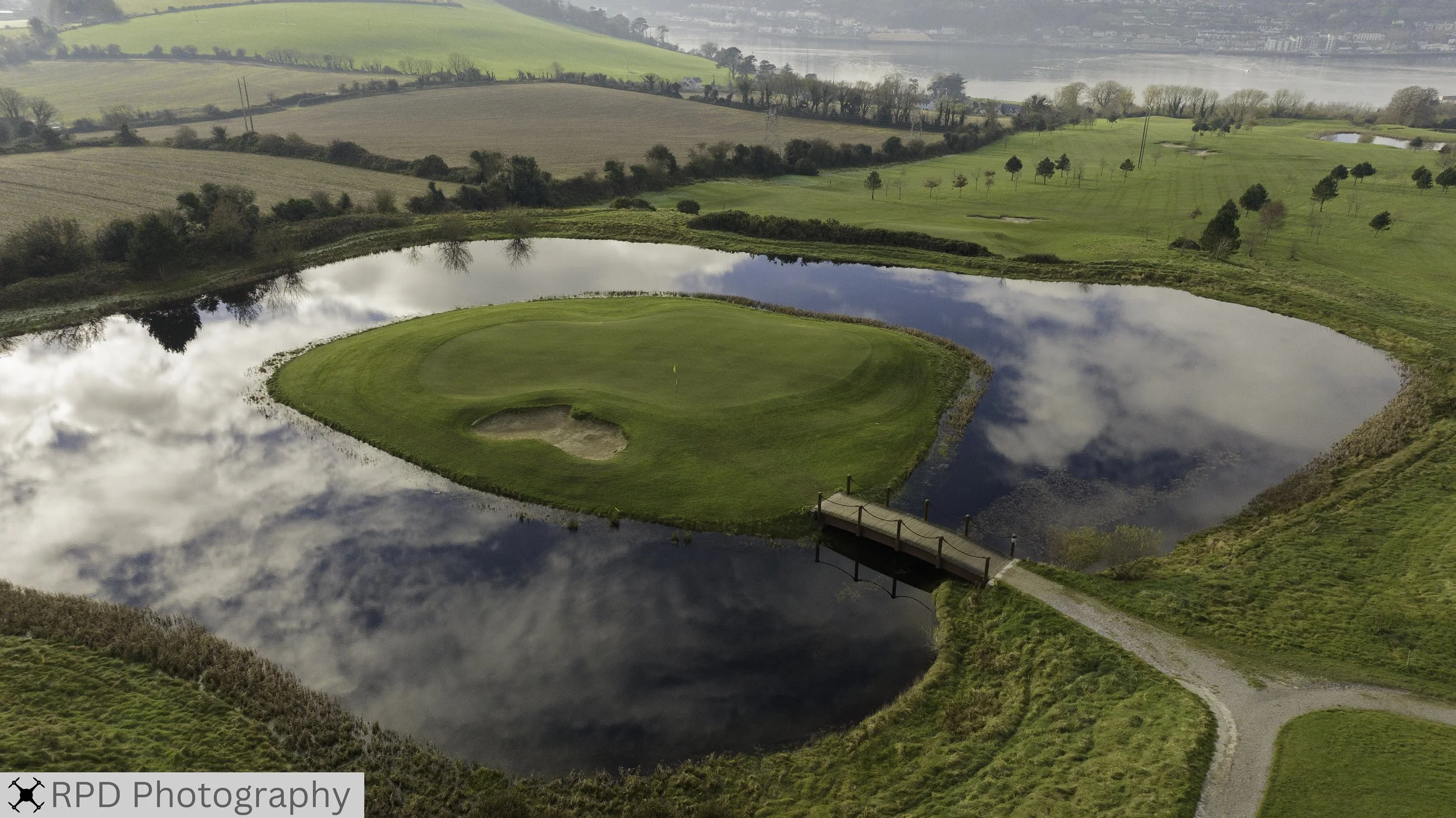 Aerial view of a golf course with a putting green surrounded by a small pond, a pathway leading to a bridge, open fields, trees, and a distant body of water under a partly cloudy sky.