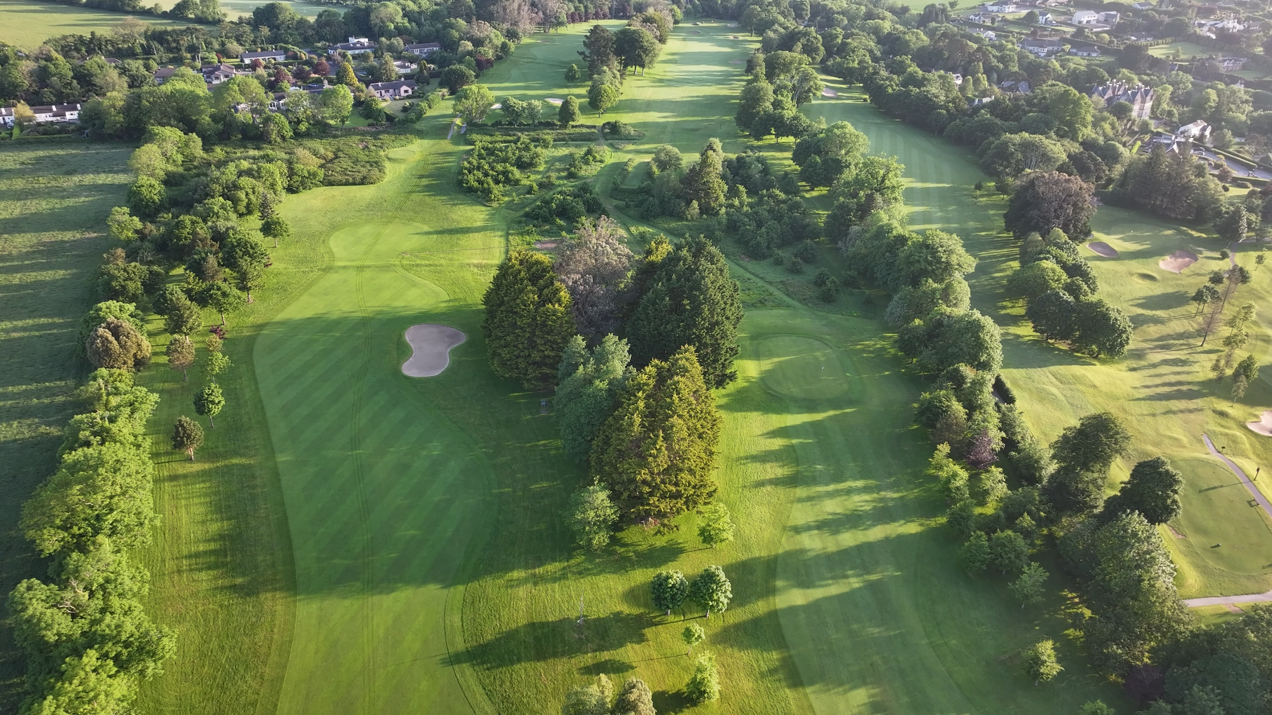 Aerial view of a golf course with green fairways, sand bunkers, and a variety of trees, surrounded by residential houses and lush greenery.
