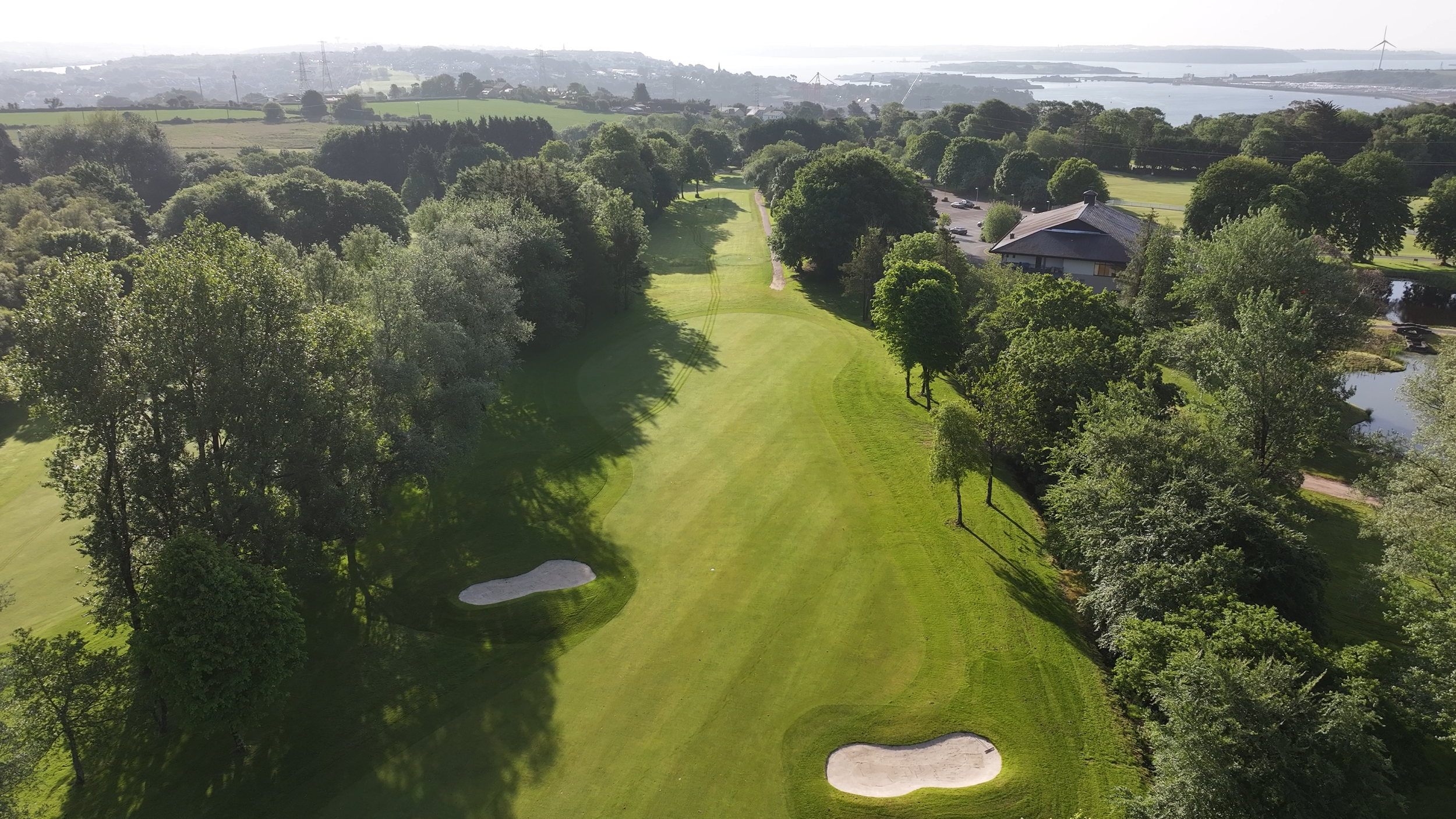 Aerial view of a golf course with lush green fairway, sand bunkers, trees on both sides, and a body of water in the background.