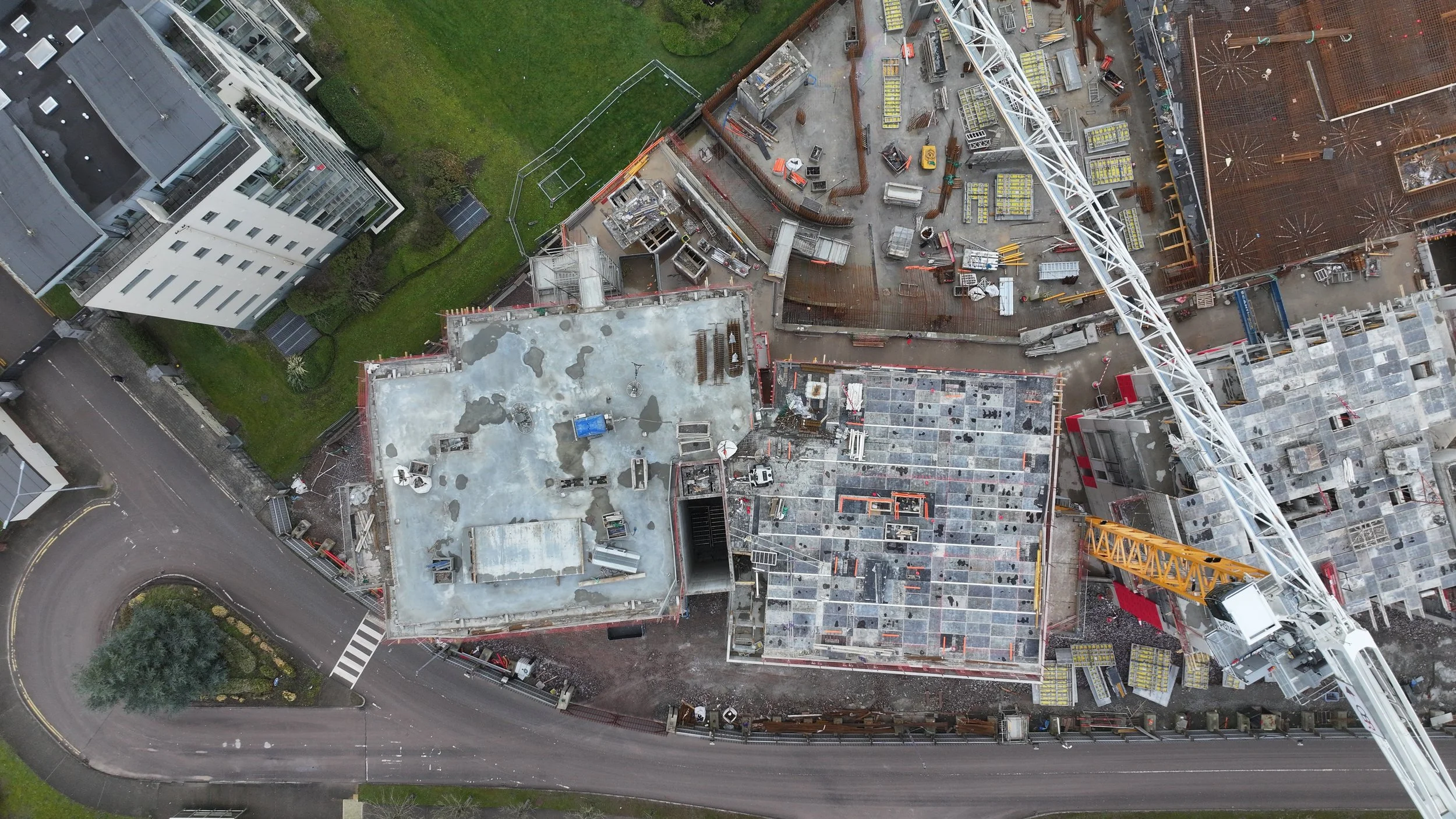 An aerial view of a construction site with cranes, building materials, and partially constructed buildings adjacent to a residential area and road.