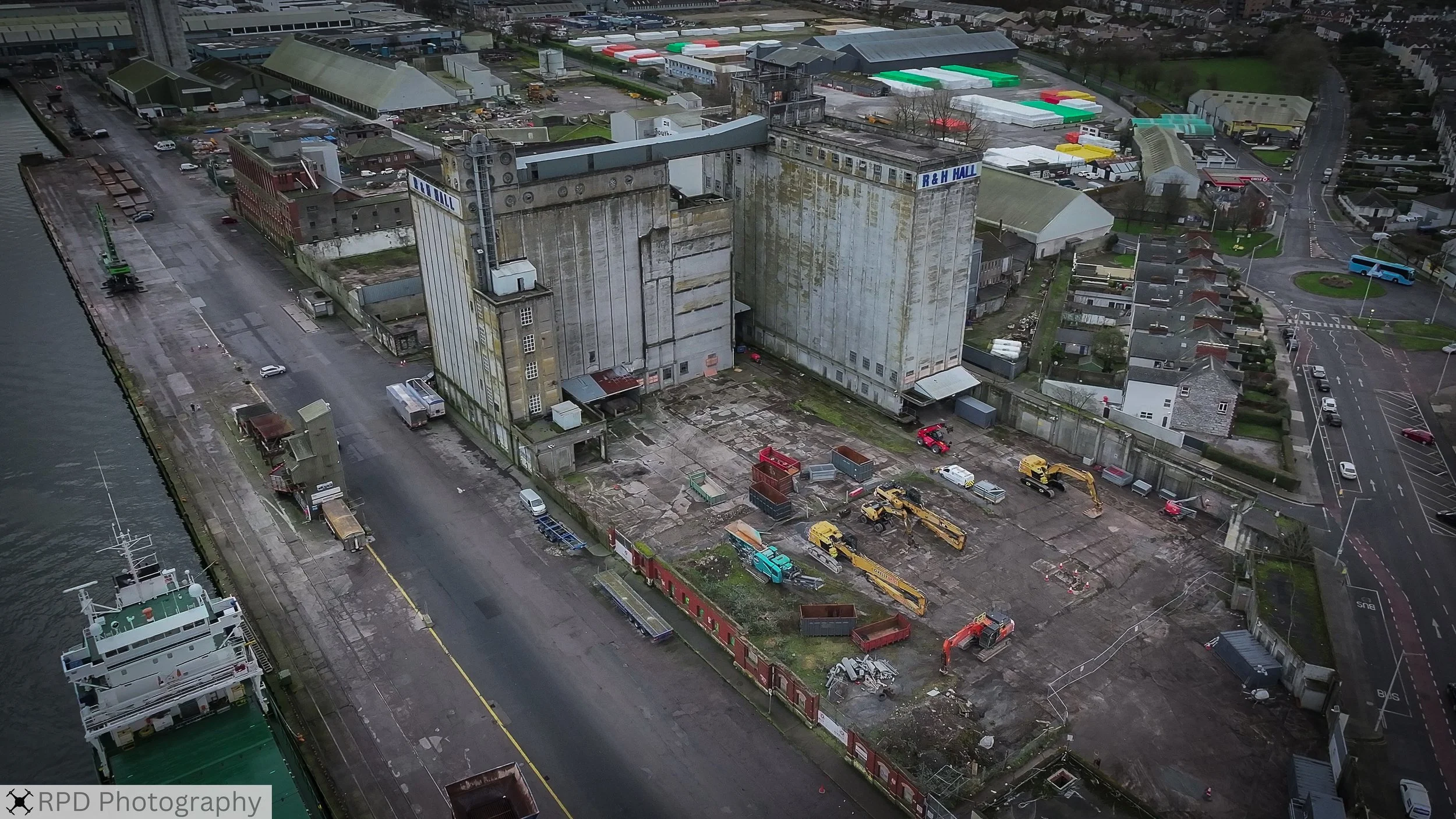 Aerial view of an industrial building next to a river, with construction vehicles and equipment in the surrounding area, and nearby streets with cars and houses.