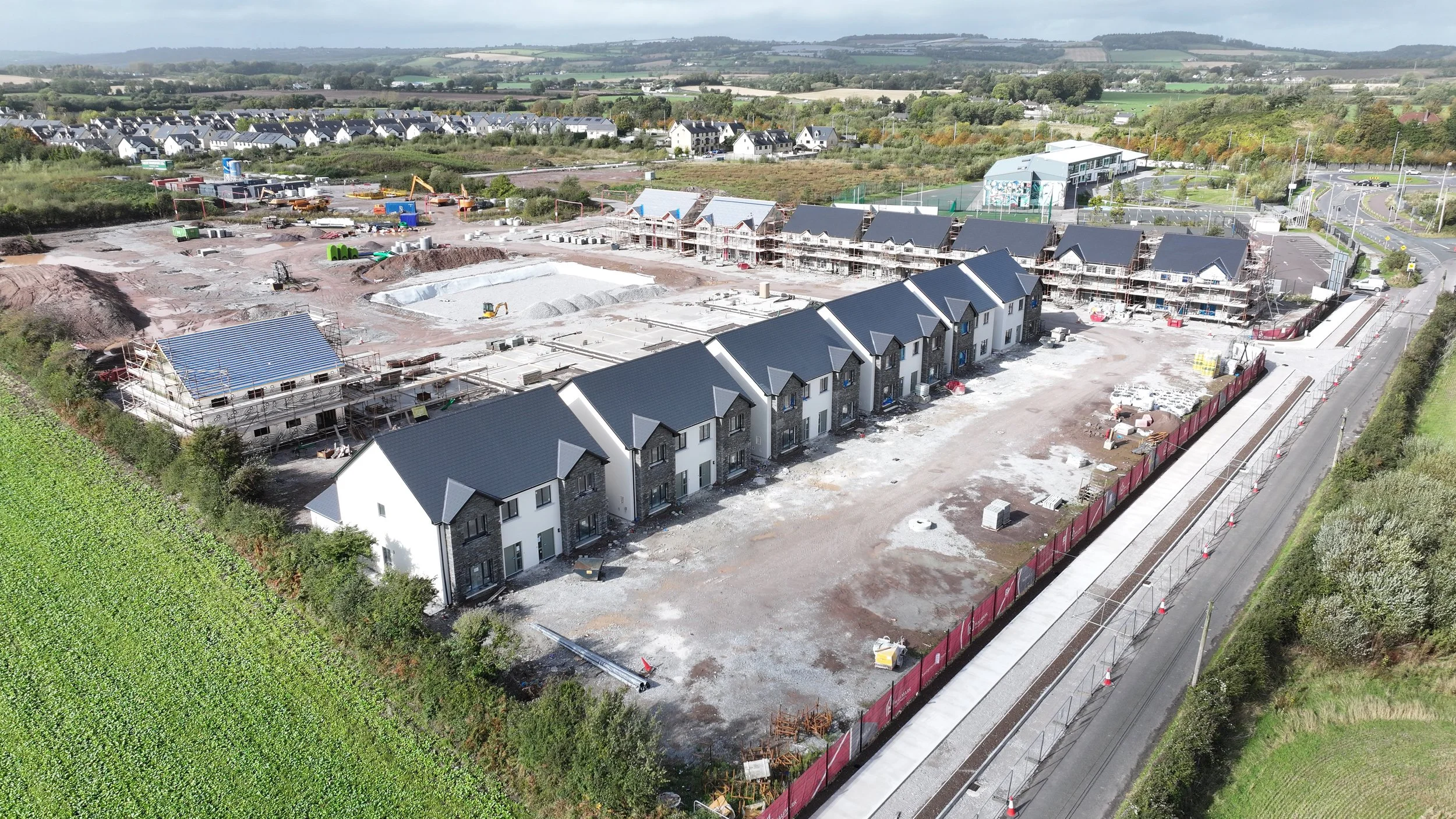 Aerial view of a construction site with new houses being built, surrounded by greenery, a road, and nearby houses in the background.