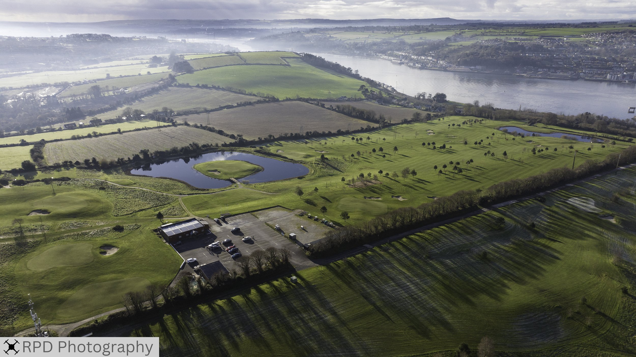 Aerial view of a golf course with green fairways, water hazards, and parking lot in the foreground, adjacent to a river with cityscape in the distance, under a cloudy sky.
