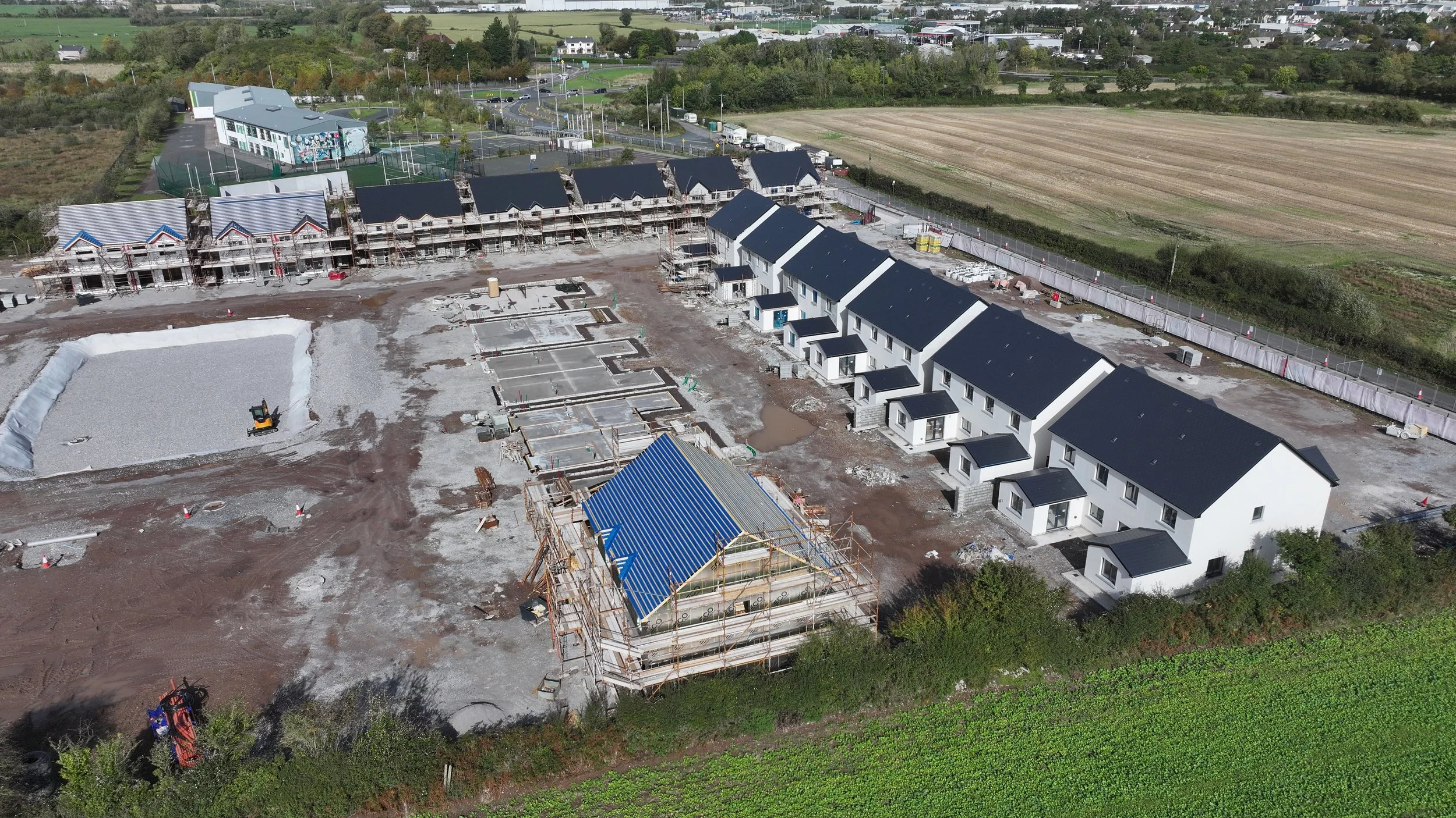 Aerial view of a construction site with multiple white houses with black roofs, some partially built, along with a small building with a blue roof. There is construction equipment, a gravel area, and nearby open fields.