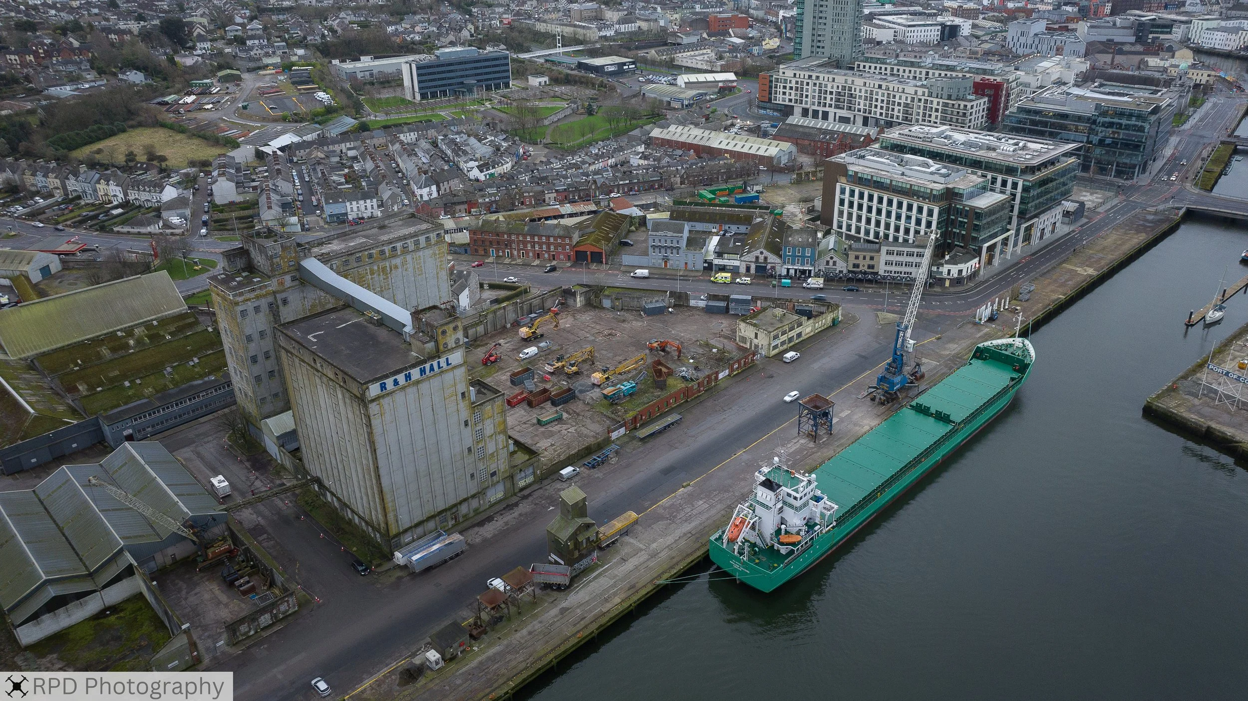 Aerial view of a harbor with a large green cargo ship docked, construction site with machinery, and city buildings in the background.