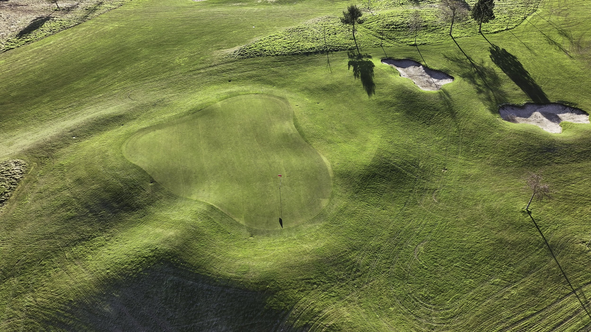 Aerial view of a golf course green with a flag, sand bunkers, and surrounding grassy landscape with trees casting shadows.