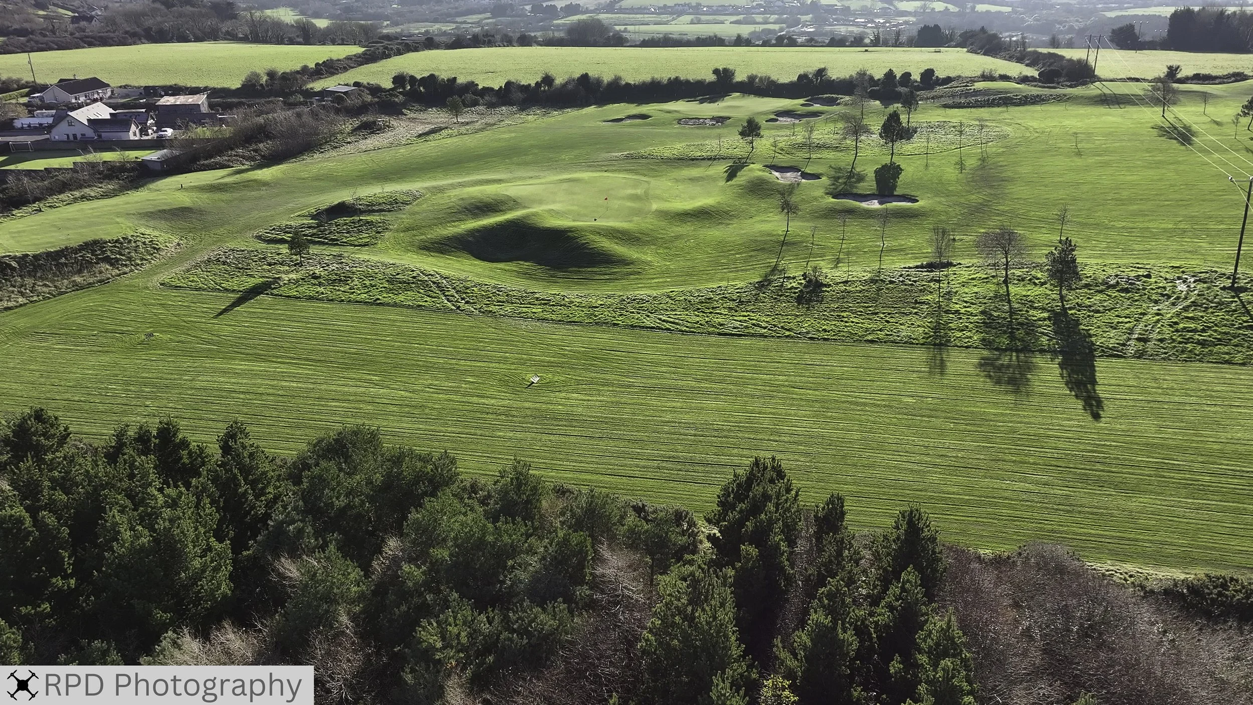 Aerial view of a golf course with lush green grass, sand traps, and small trees, surrounded by houses and open fields.