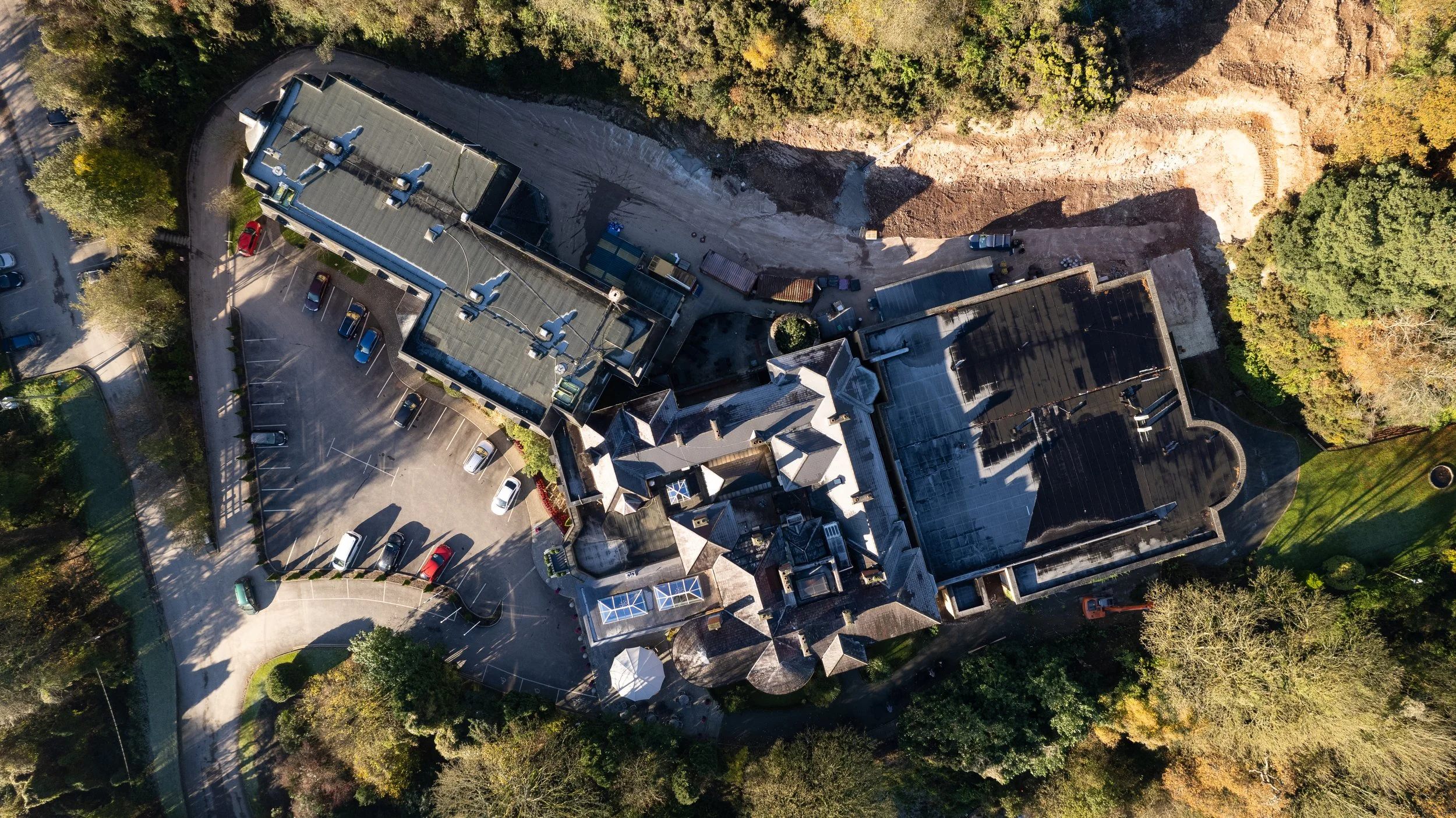 Aerial view of a large building complex surrounded by trees, parking lot with parked cars, and a nearby construction area.