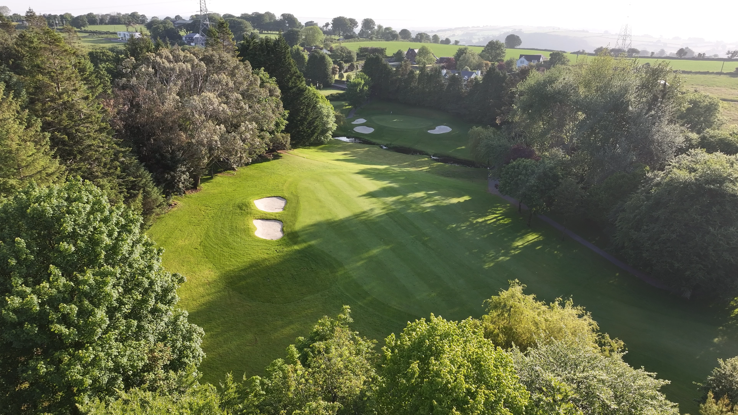 An aerial view of a golf course with green fairways, sand bunkers, surrounded by trees and some residential houses in the background.
