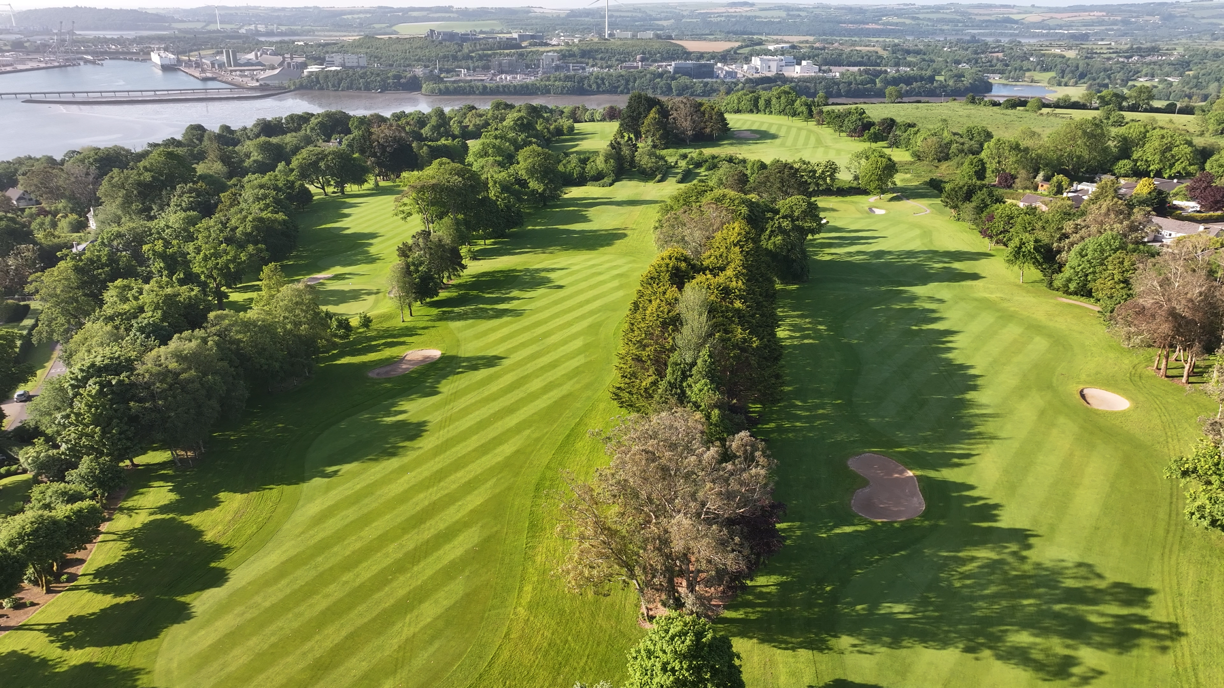 Aerial view of a golf course with neatly mowed fairways, sand bunkers, and trees, near a river and industrial area in the background.