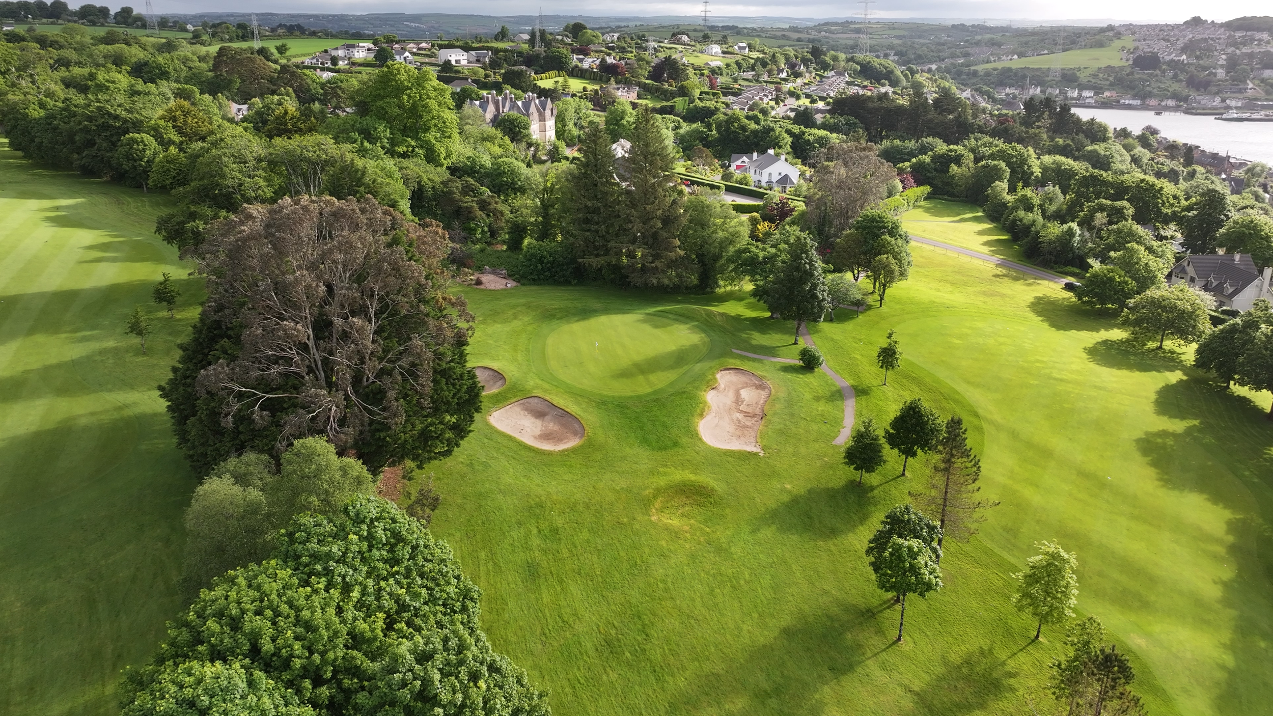 Aerial view of a golf course surrounded by green trees and a lush landscape, with residential houses and a water body visible in the background.