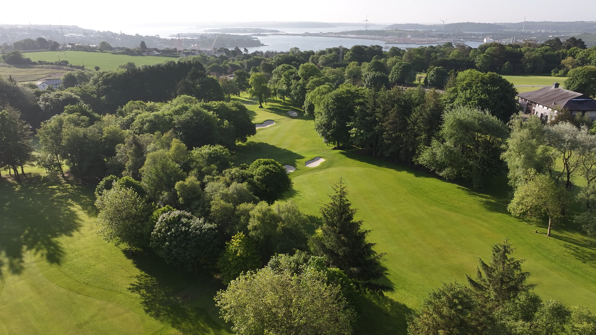 Aerial view of a lush green golf course with sand traps, surrounded by numerous trees, and a body of water in the background.