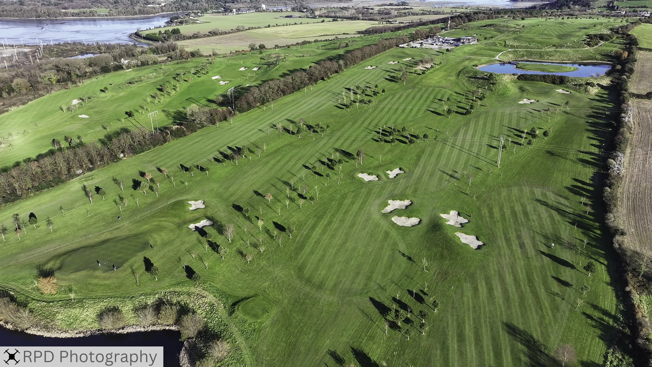 Aerial view of a lush green golf course with multiple sand traps, trees, and water hazards, surrounded by fields and a river in the background.