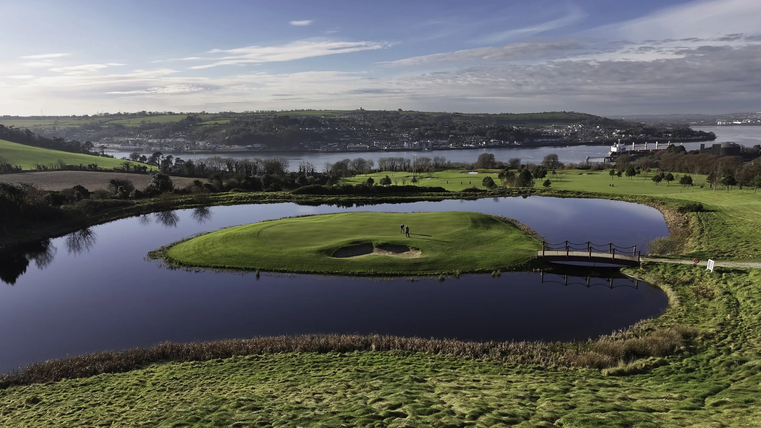 A scenic golf course with a pond in the foreground, a bridge over the pond, and a couple standing on the green. There is a large body of water and a cityscape with industrial buildings and smokestacks in the background under a partly cloudy sky.