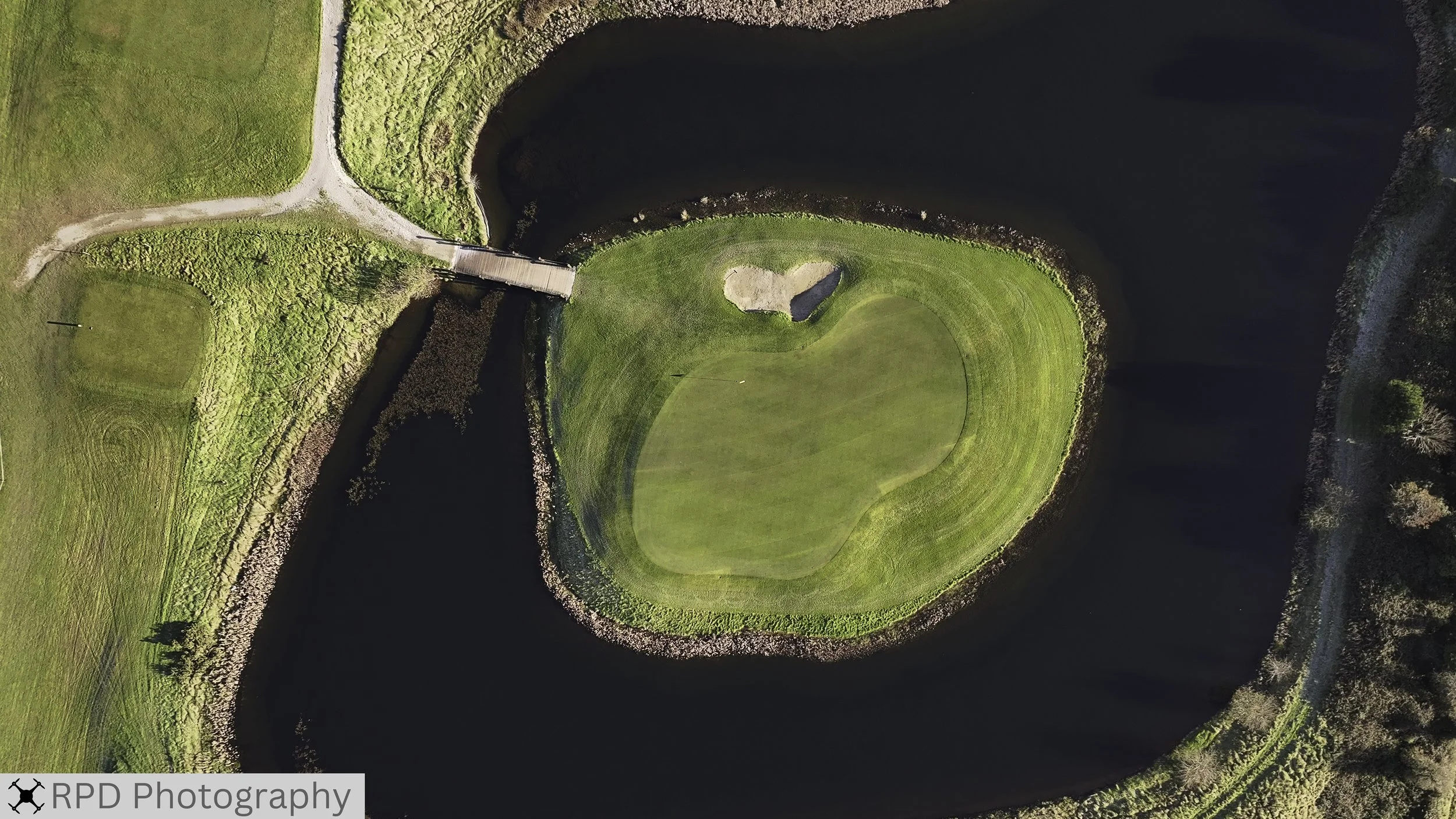 An aerial view of a golf course green surrounded by water, with a small sand trap near the center and a bridge connecting two areas of the course.
