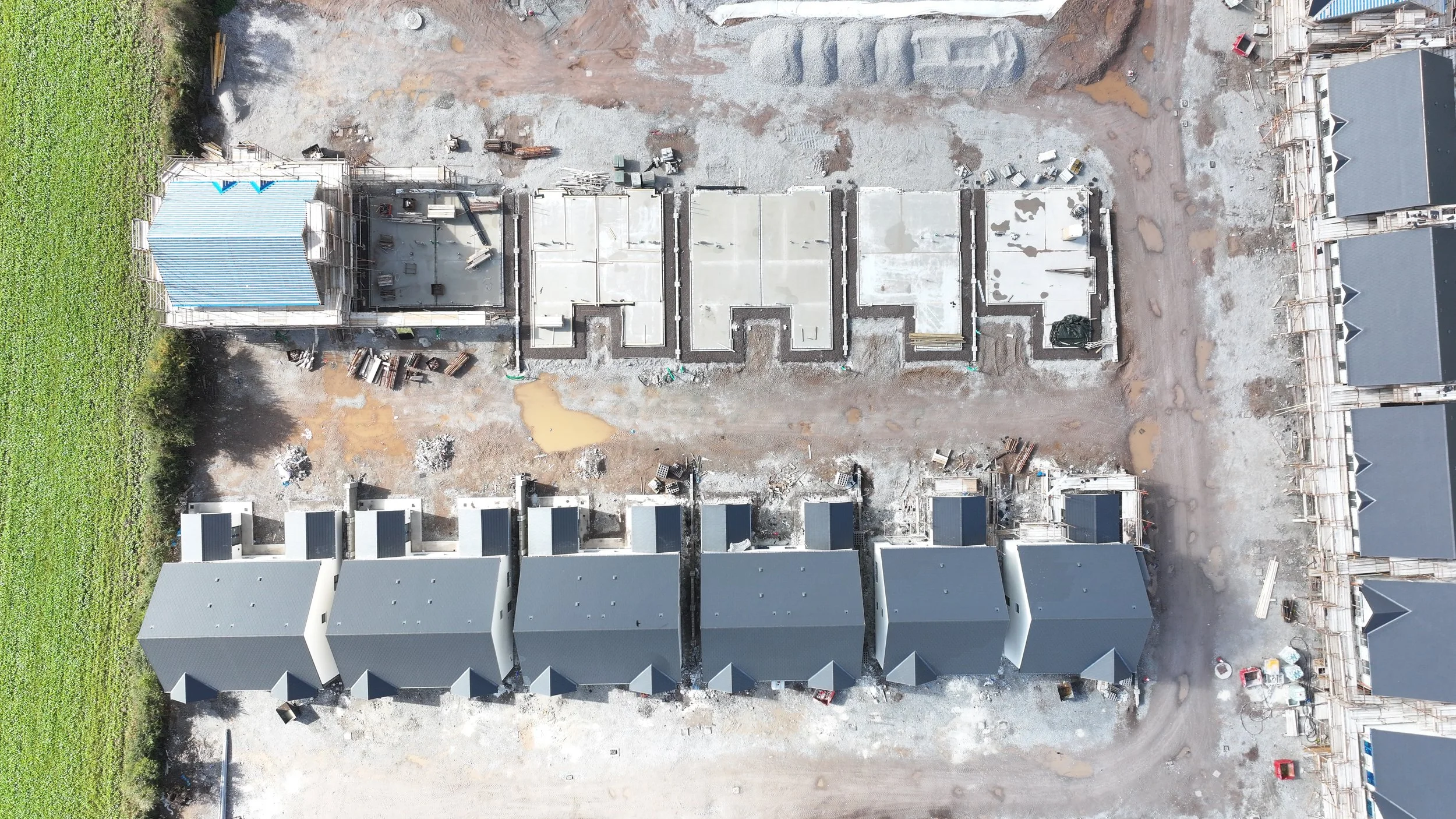 An aerial view of a construction site showing multiple partially built houses with gray roofs and the foundation of another house under construction, surrounded by dirt, construction materials, and equipment.